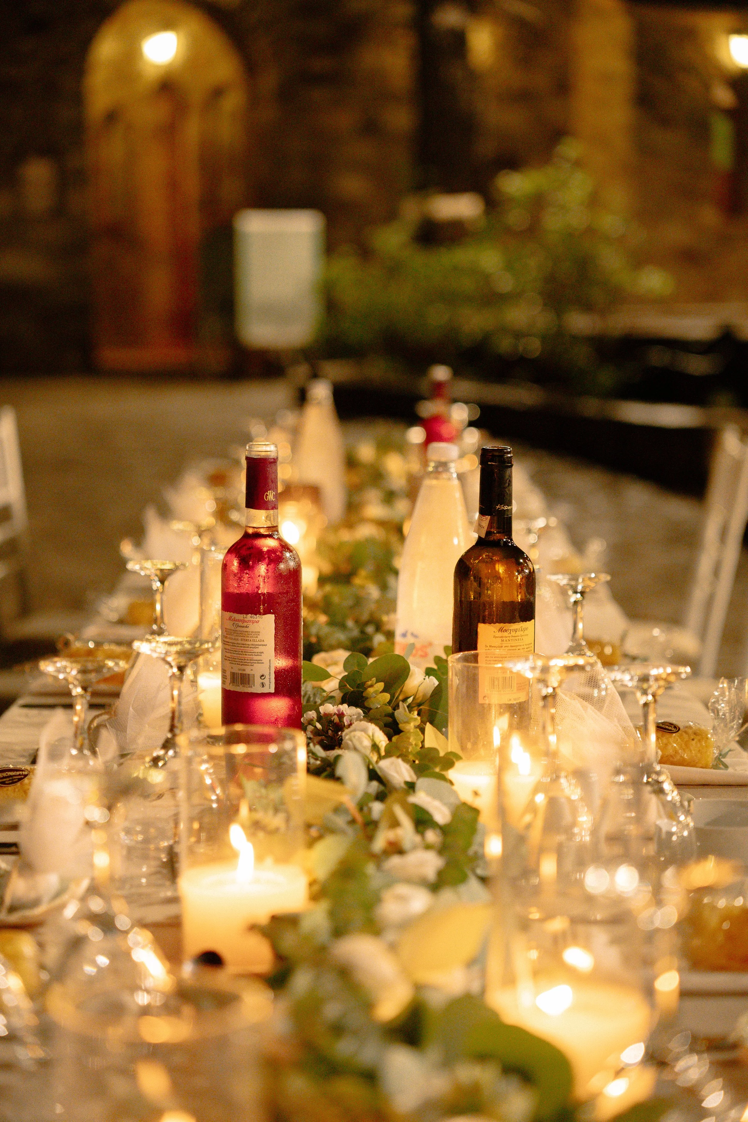 A long dining table set for an event with wine bottles, candles, and floral decorations in a warmly lit outdoor setting at night.