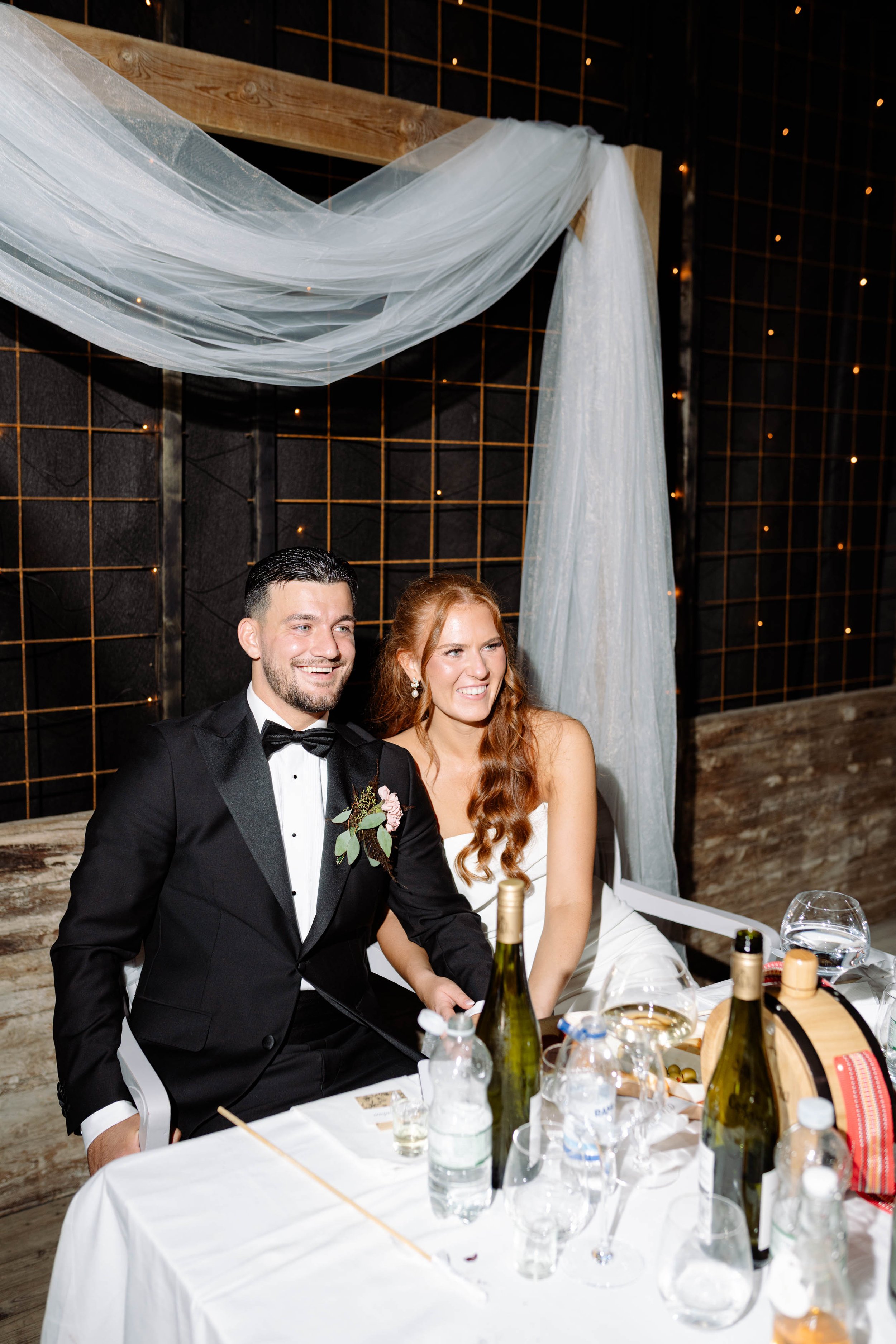 A newlywed couple sitting at a table during their wedding reception, smiling happily. The groom is dressed in a black tuxedo with a boutonnière, and the bride is wearing a white wedding dress. The table has bottles of wine, water, and glasses, with a
