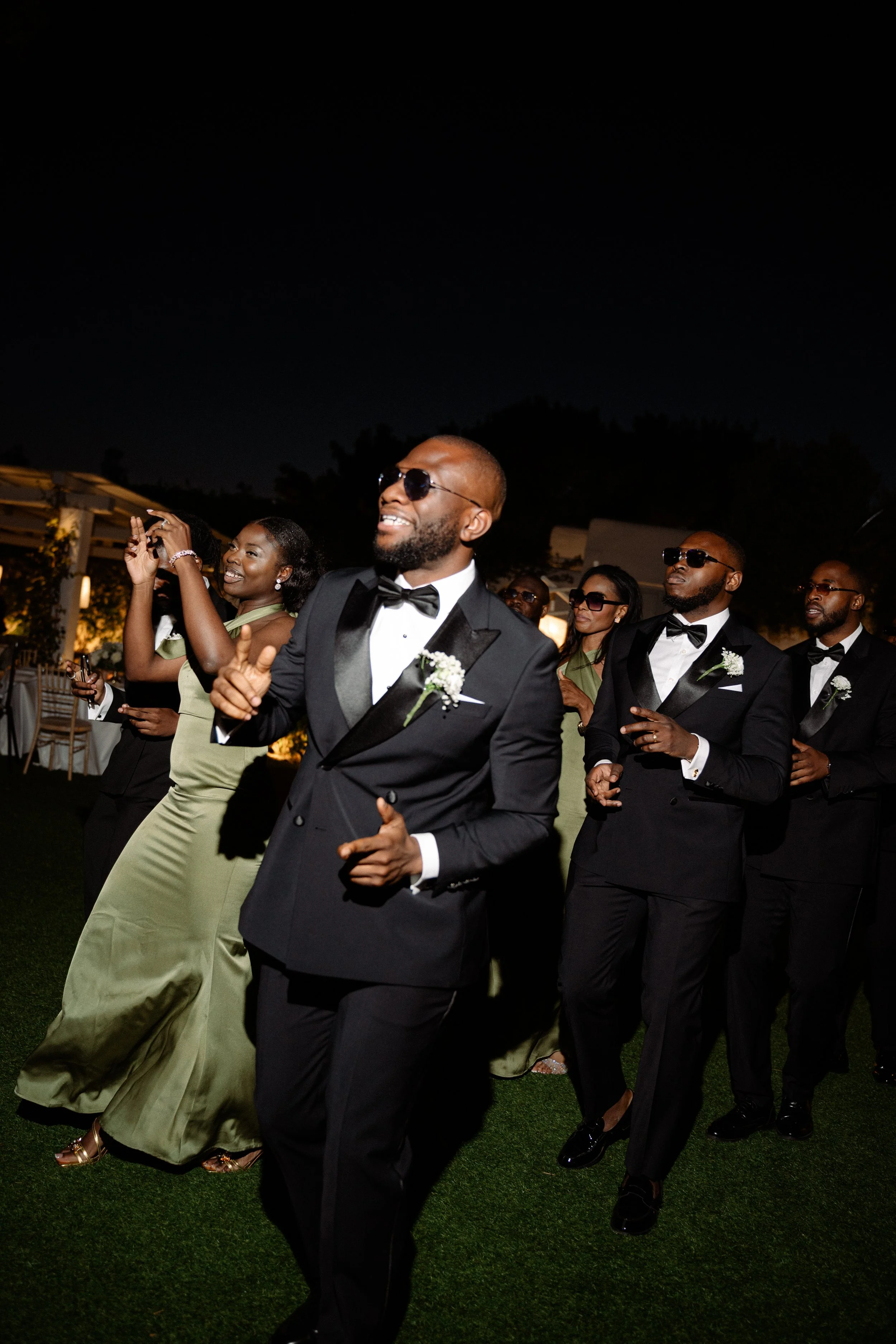 Group of people dressed in formal attire dancing outdoors at night, some wearing sunglasses, smiling and enjoying themselves.