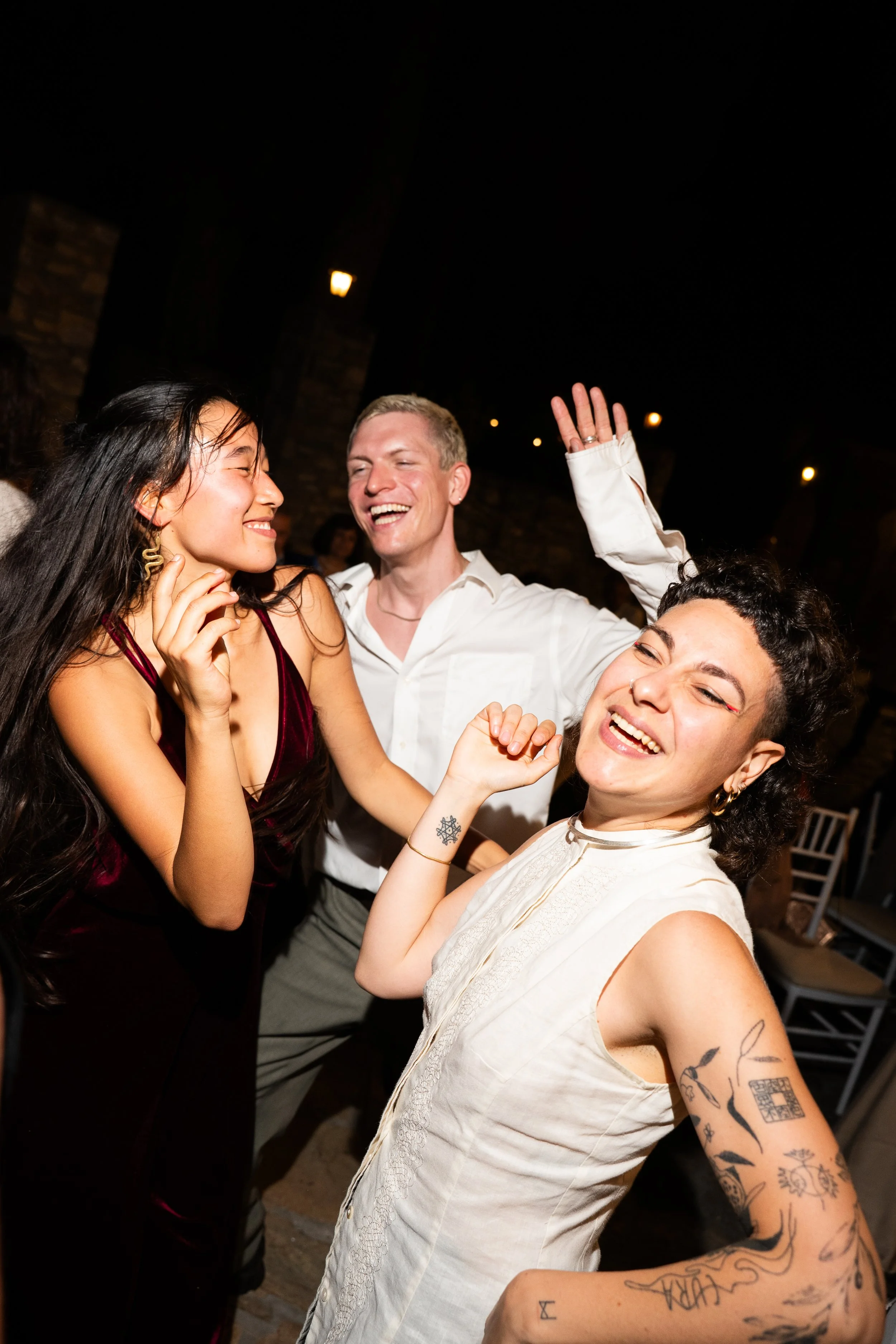 Three people dancing and smiling at a party or celebration at night, with some seated guests and stone wall background.