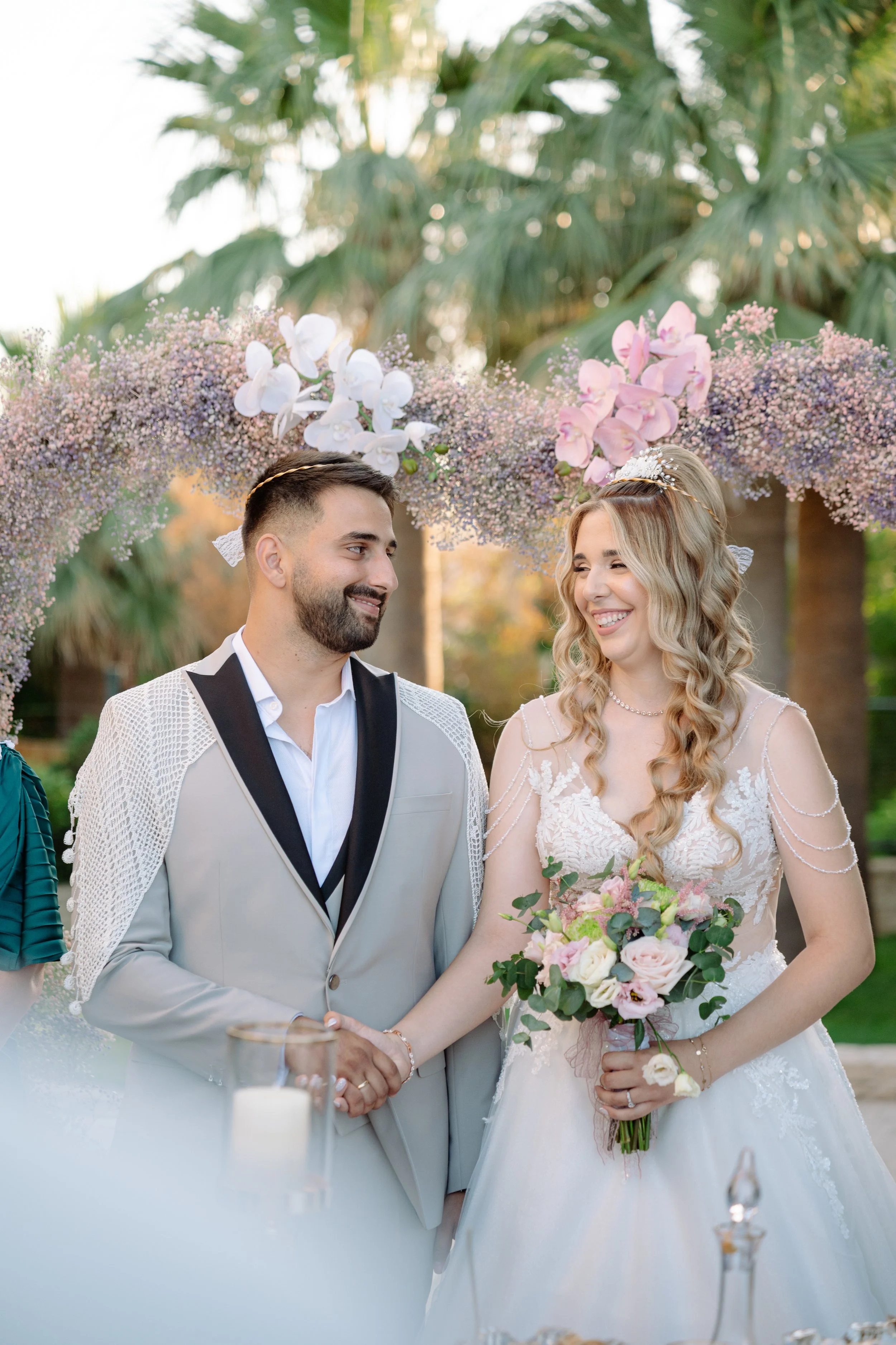 A bride and groom smiling and holding hands during their wedding ceremony outdoors, with floral decorations in the background.
