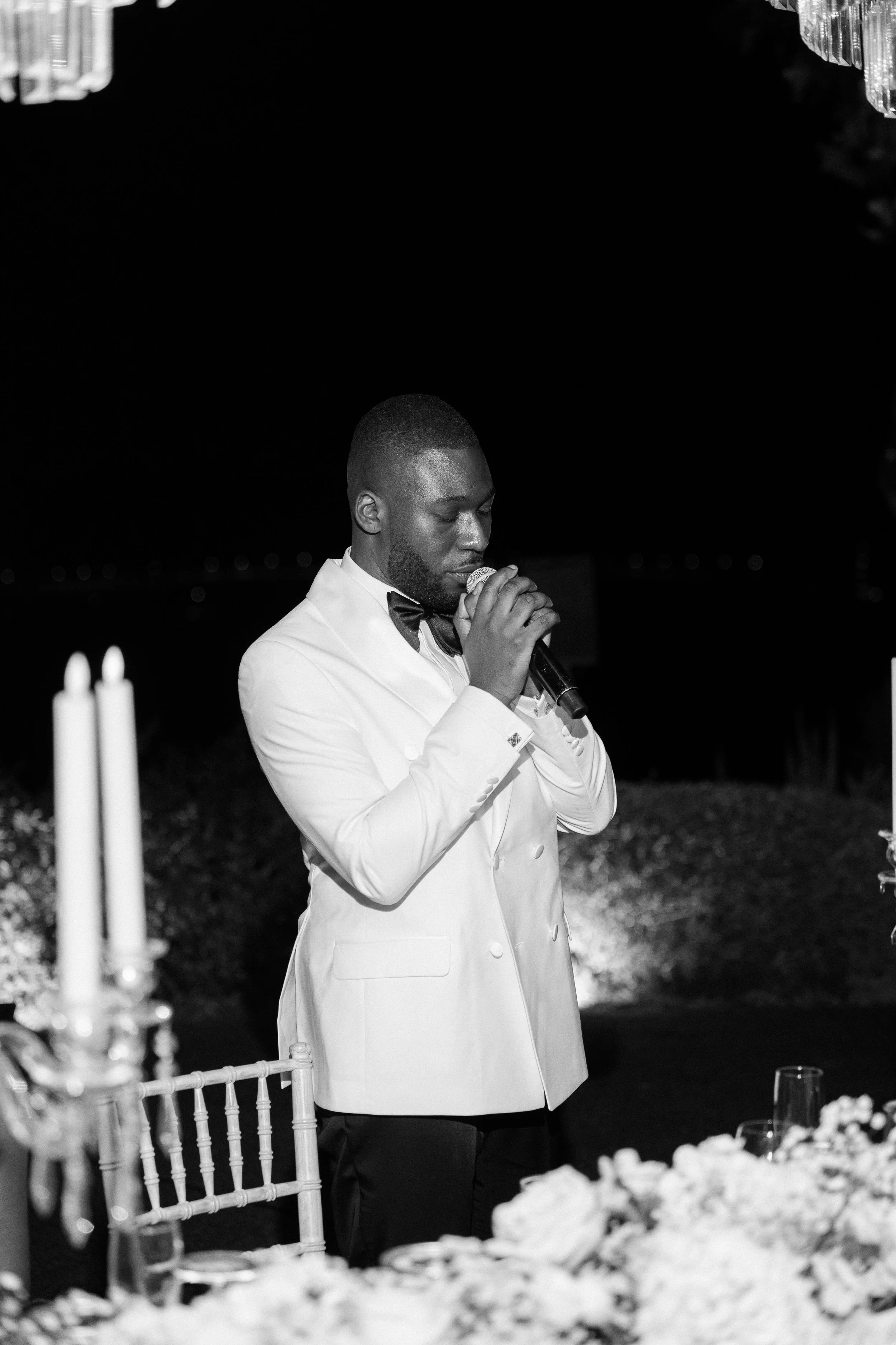 A man in a white tuxedo jacket with a bow tie, holding a microphone and speaking at a formal event, with a table decorated with flowers and candles in the foreground.