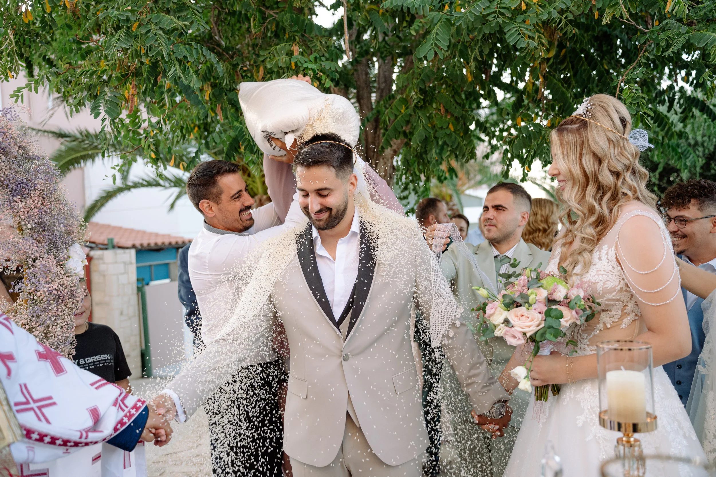 A wedding ceremony outdoors with a bride and groom surrounded by friends and family, as rice is thrown over the couple.