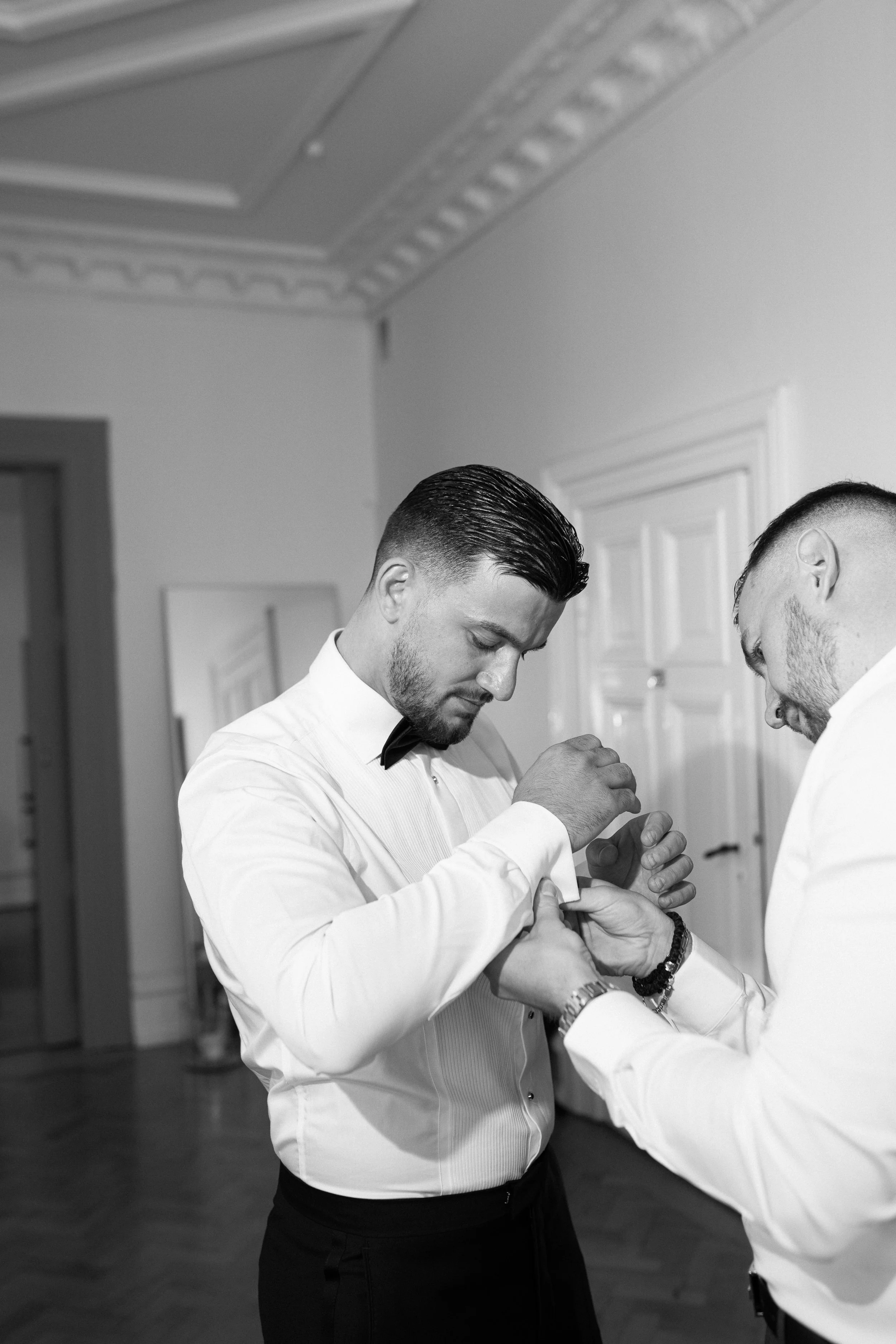Two men in formal attire, one helping the other with cufflinks, inside a room with ornate ceiling molding and a mirror.