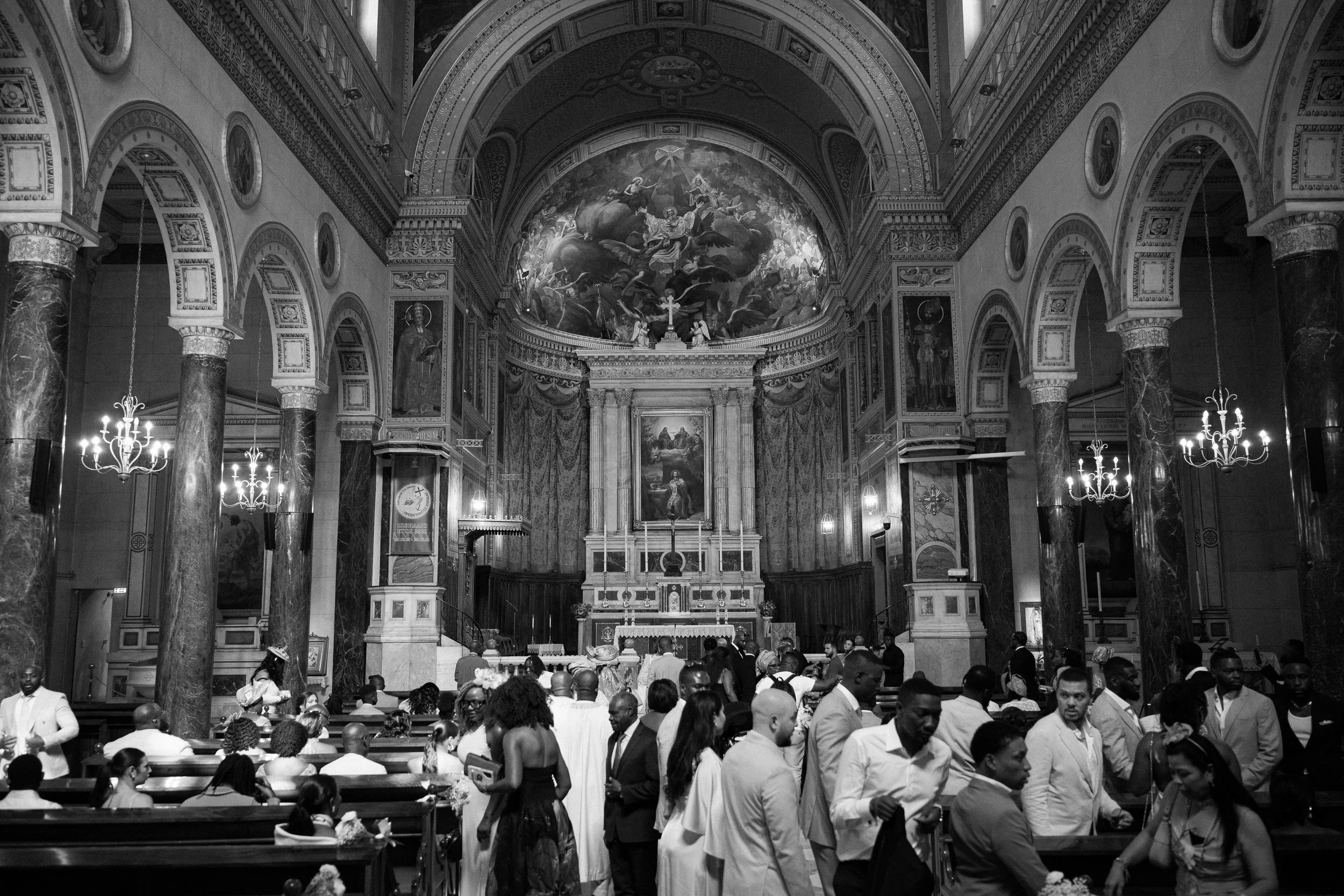A black-and-white photo of a church interior filled with wedding guests, with an altar in the background, ornate columns, and a painted ceiling.