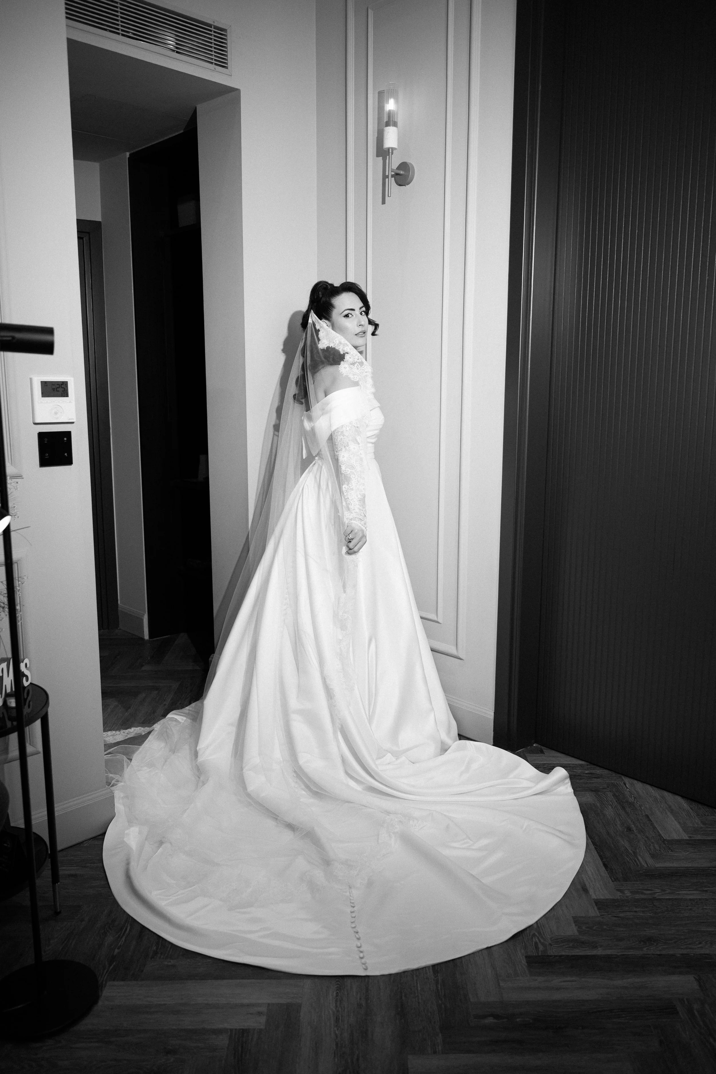 A woman in a wedding dress with a long train and veil standing indoors next to a wall with modern light fixture. In black and white.