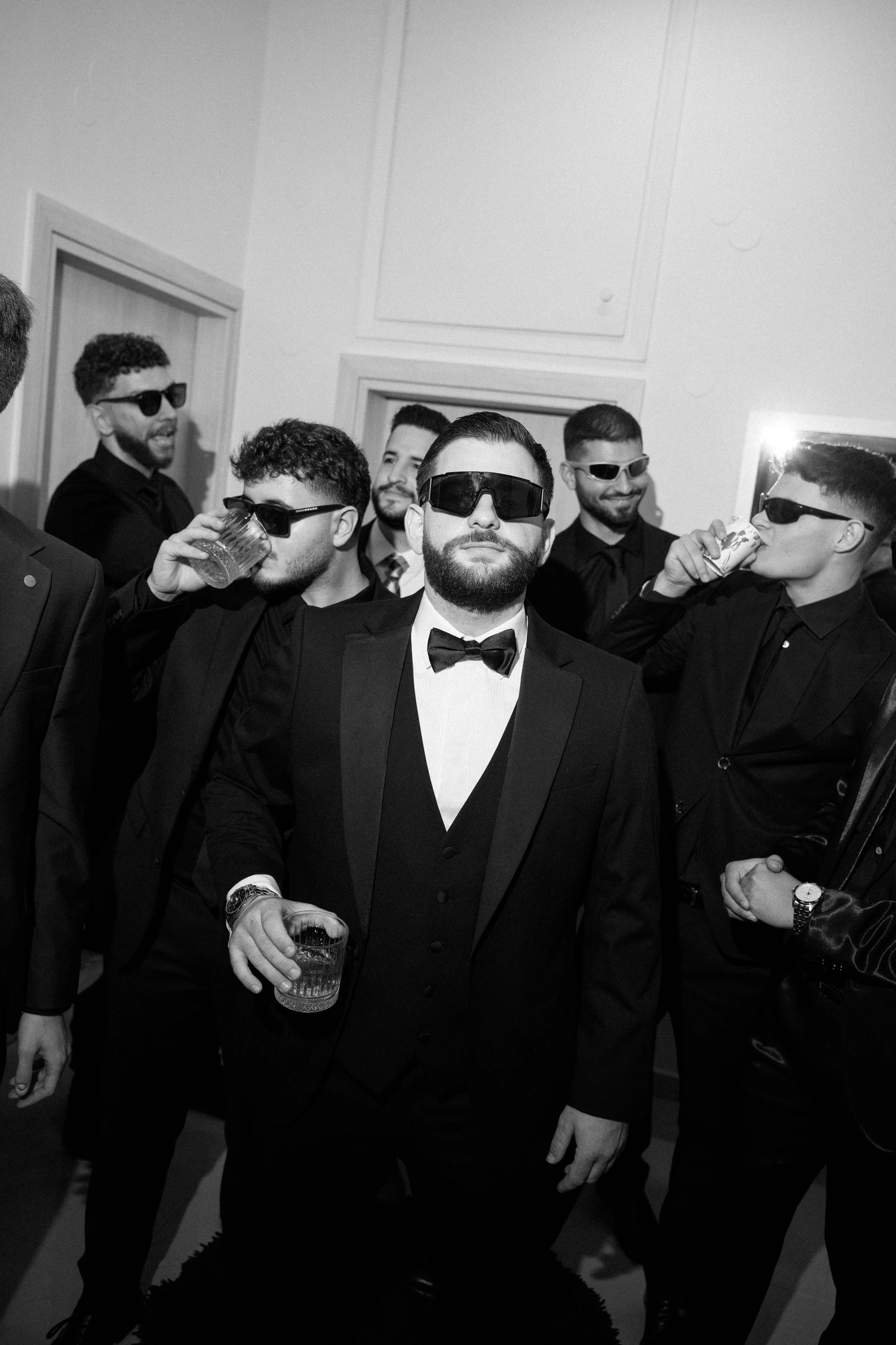 Group of men at a party wearing tuxedos and sunglasses, some holding drinks, in a black-and-white photo.