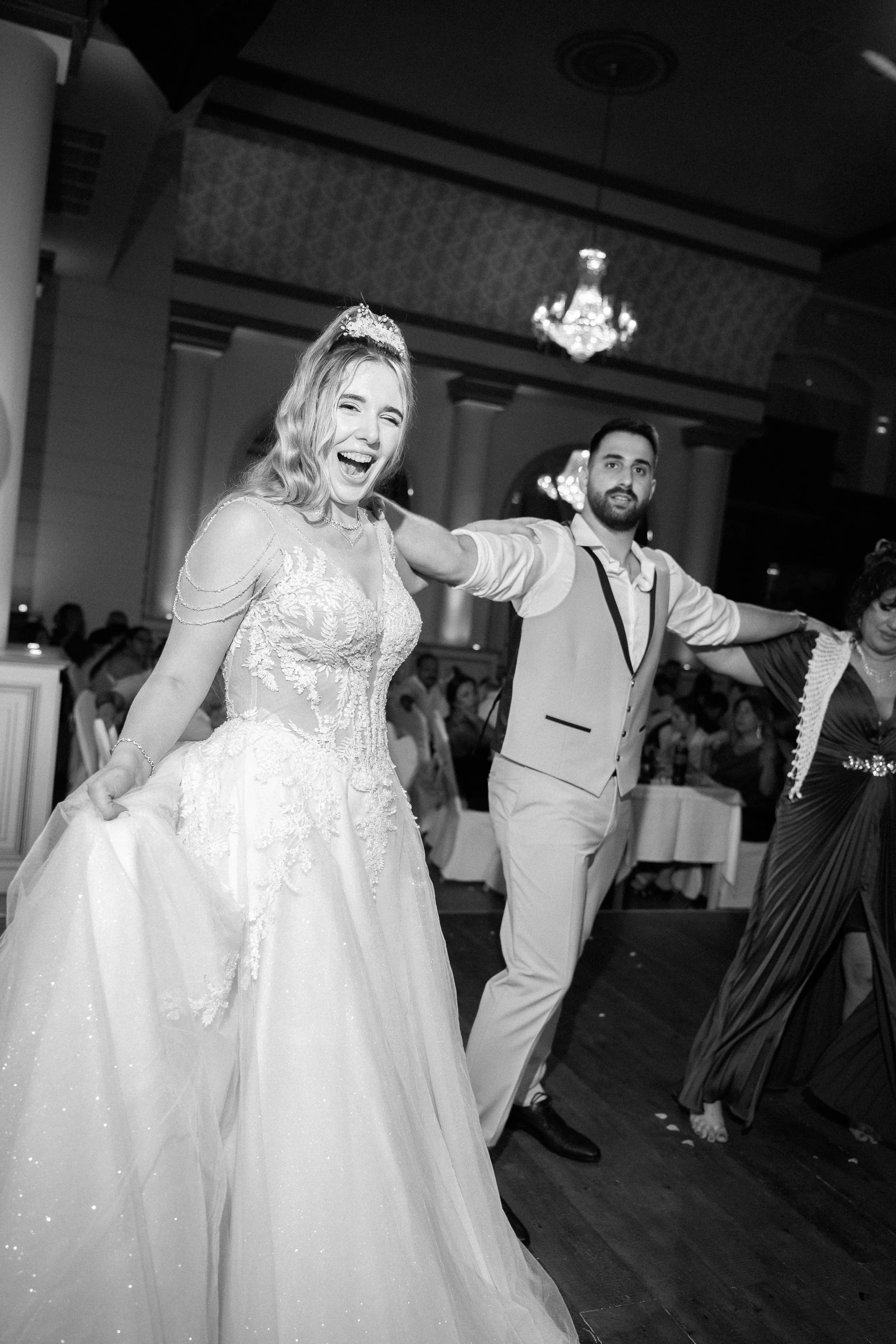 A bride in a wedding dress with lace detail and a tiara, smiling and holding up her gown, at a wedding reception with guests in the background. A man in a vest and shirt, with his arms outstretched, is standing next to her.