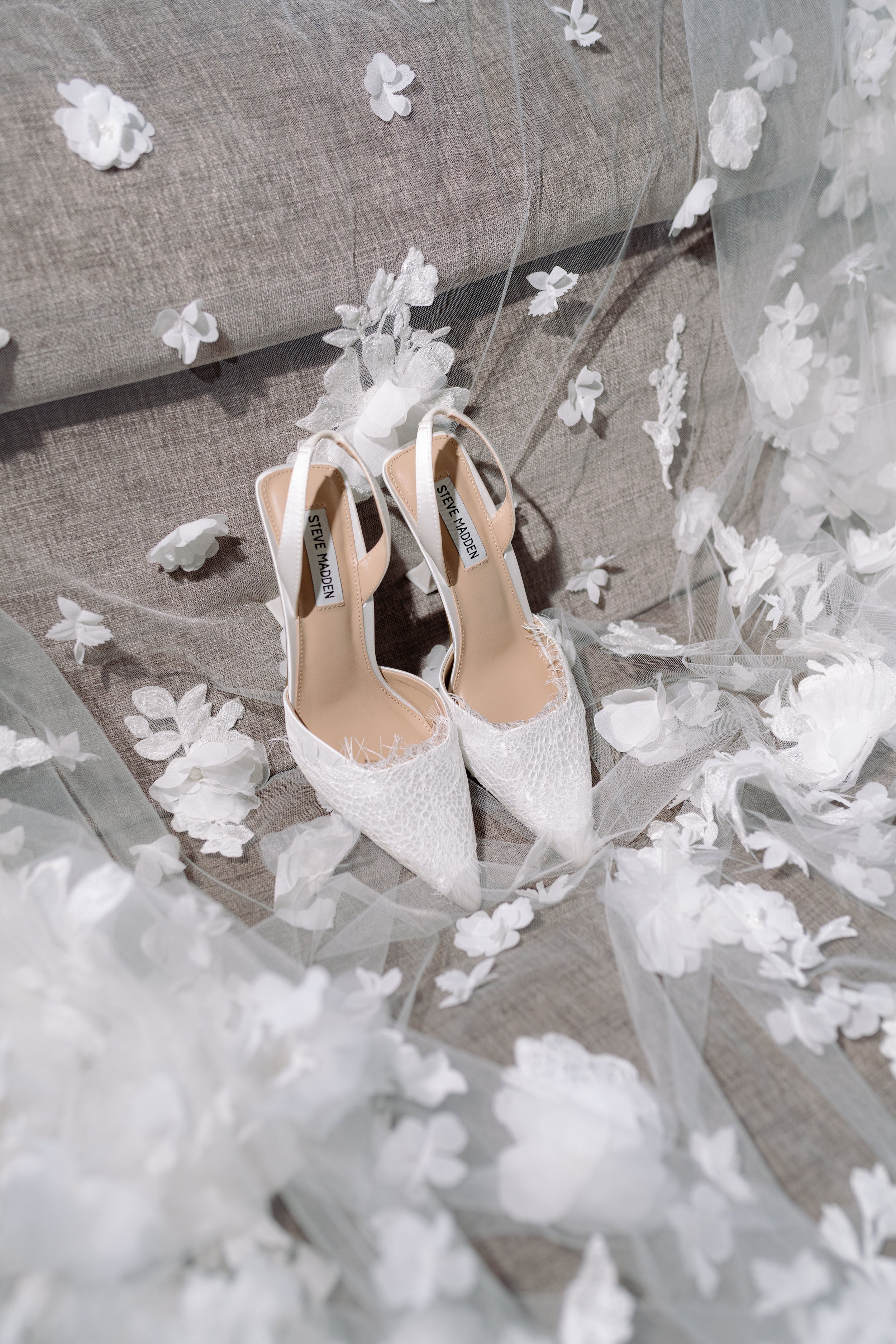 White pointed-toe high heels with lace details, placed on a gray couch surrounded by white flower petals and tulle, with visible tags labeled 'Steve Madden' inside the shoes.