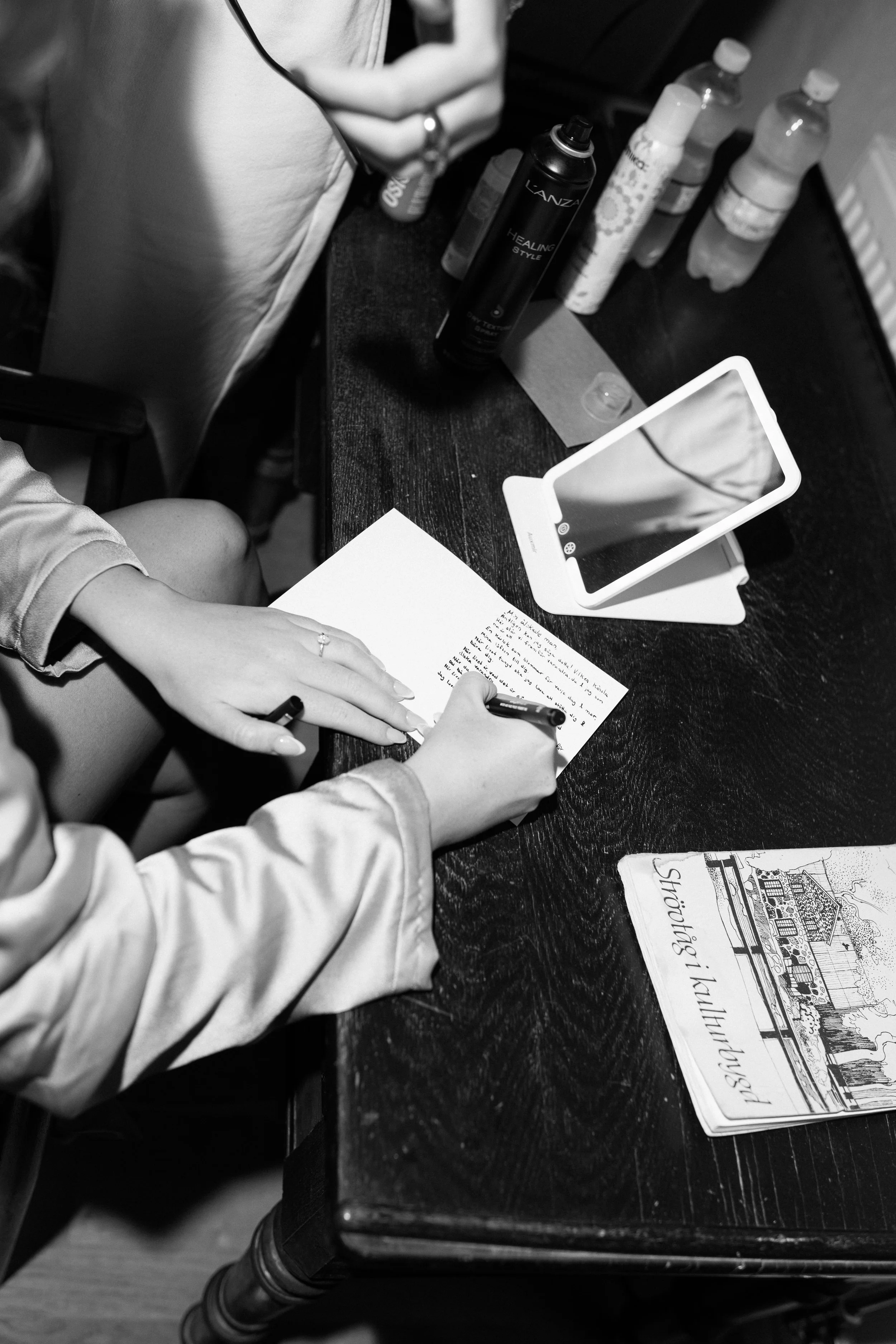 A person writing on a piece of paper at a dark wooden table. On the table are various bottles, a phone propped up on a white stand, and a brochure or newspaper with a sketch of a house and the text 'Sharing'.