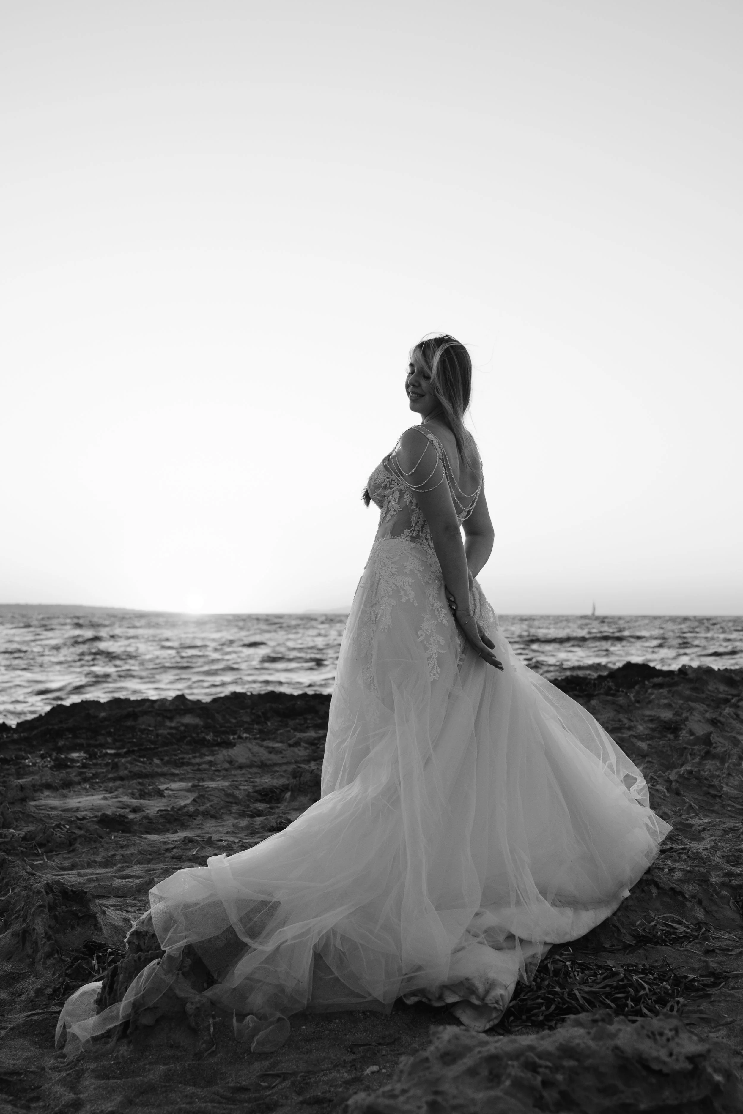 A woman in a wedding dress standing on rocky terrain by the ocean during sunset, smiling with her eyes closed.