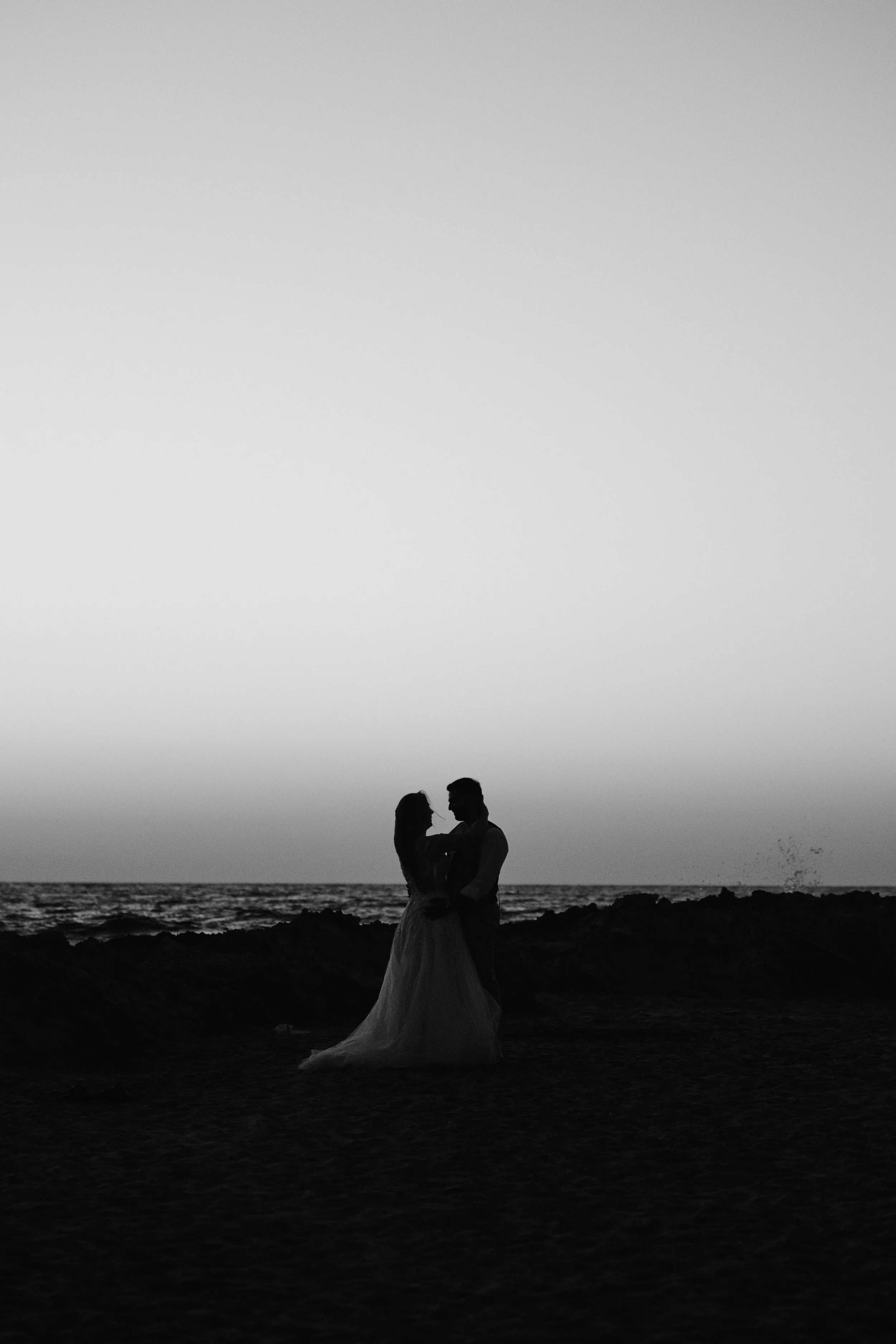 Silhouette of a couple standing on the beach, holding each other, against a cloudy sky and ocean background, during sunset or sunrise.