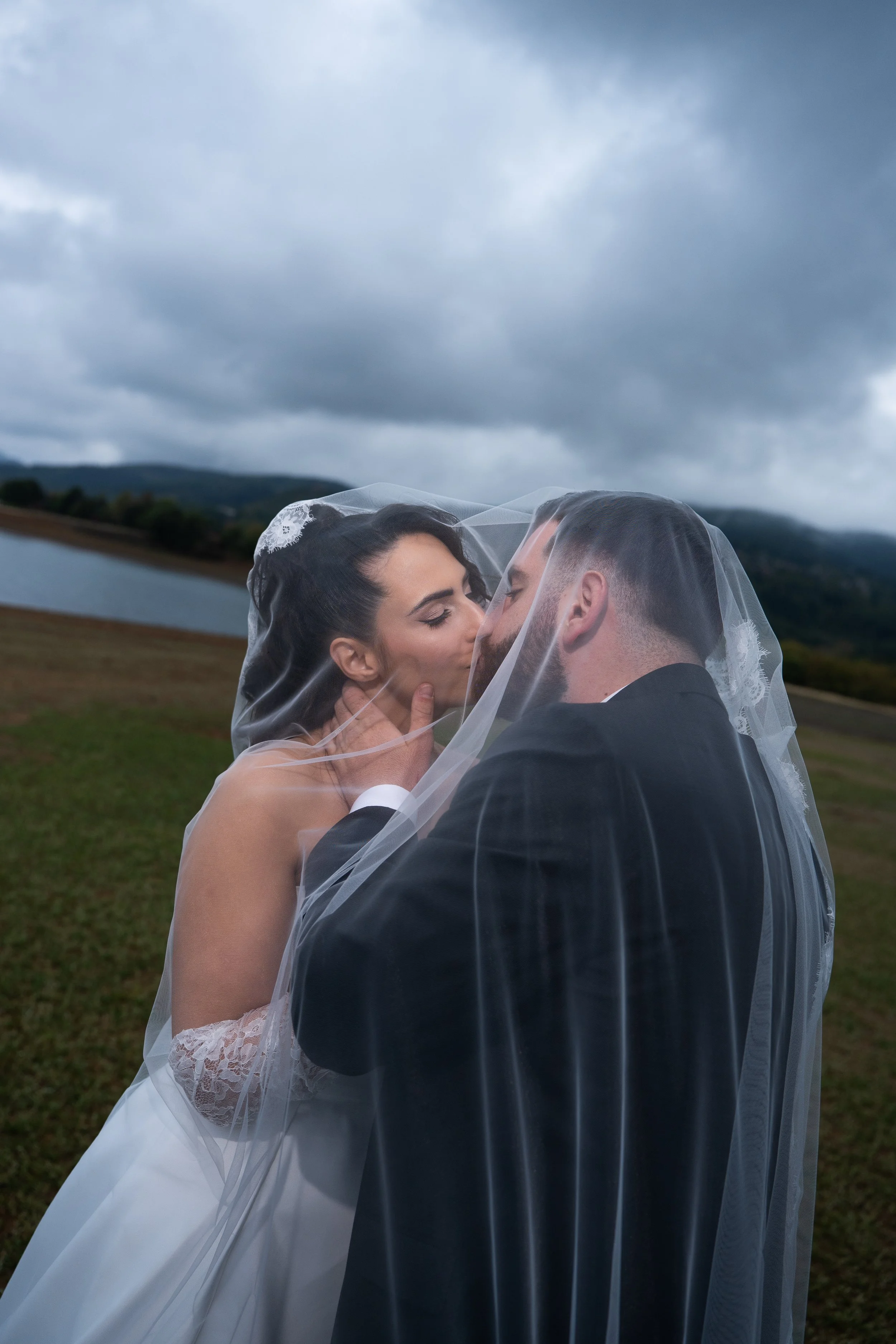 Bride and groom kissing outdoors under a veil with cloudy sky and lake in background.