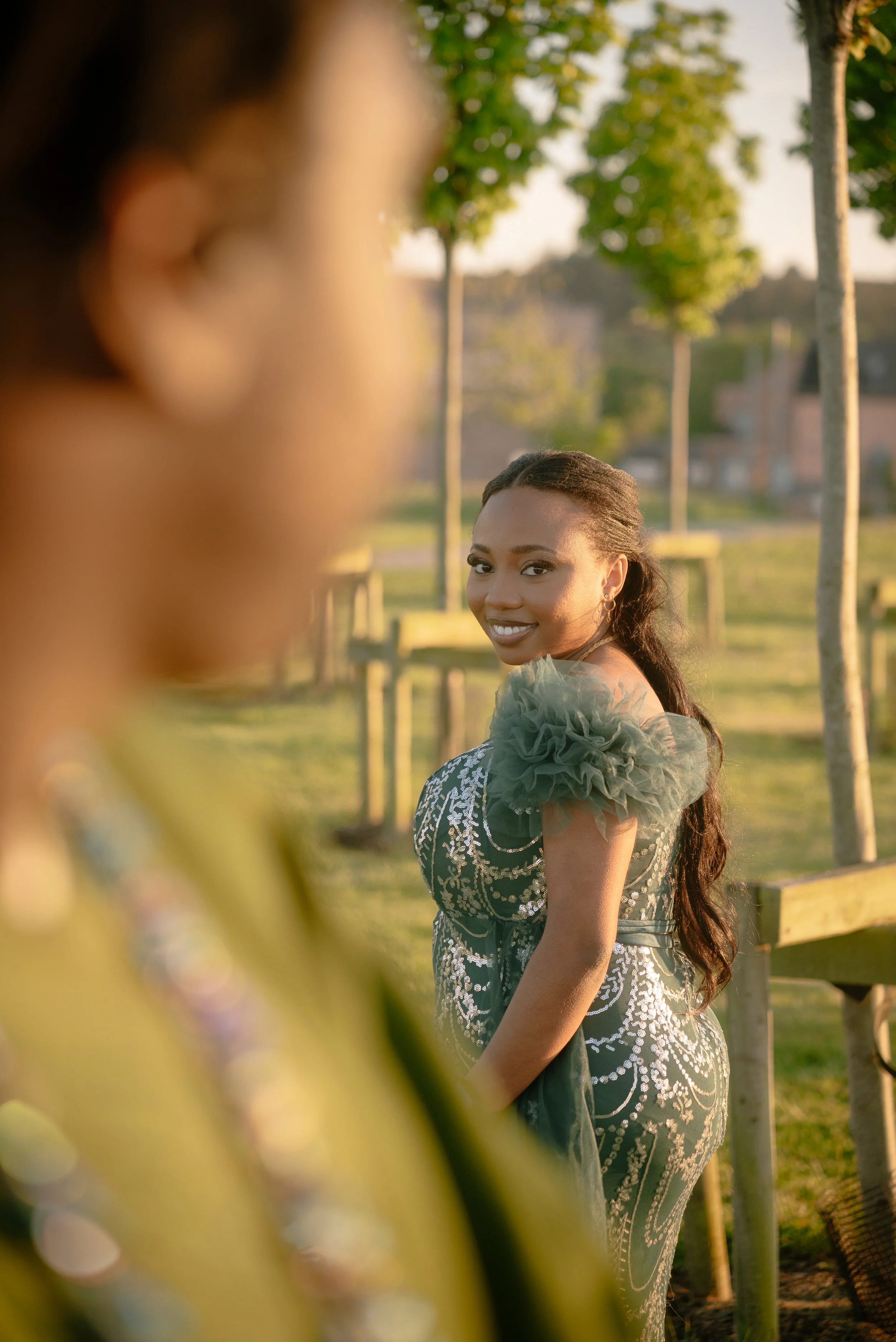 A woman smiling and looking over her shoulder outdoors, with trees and a wooden fence in the background, wearing a dark dress with tulle shoulder details.