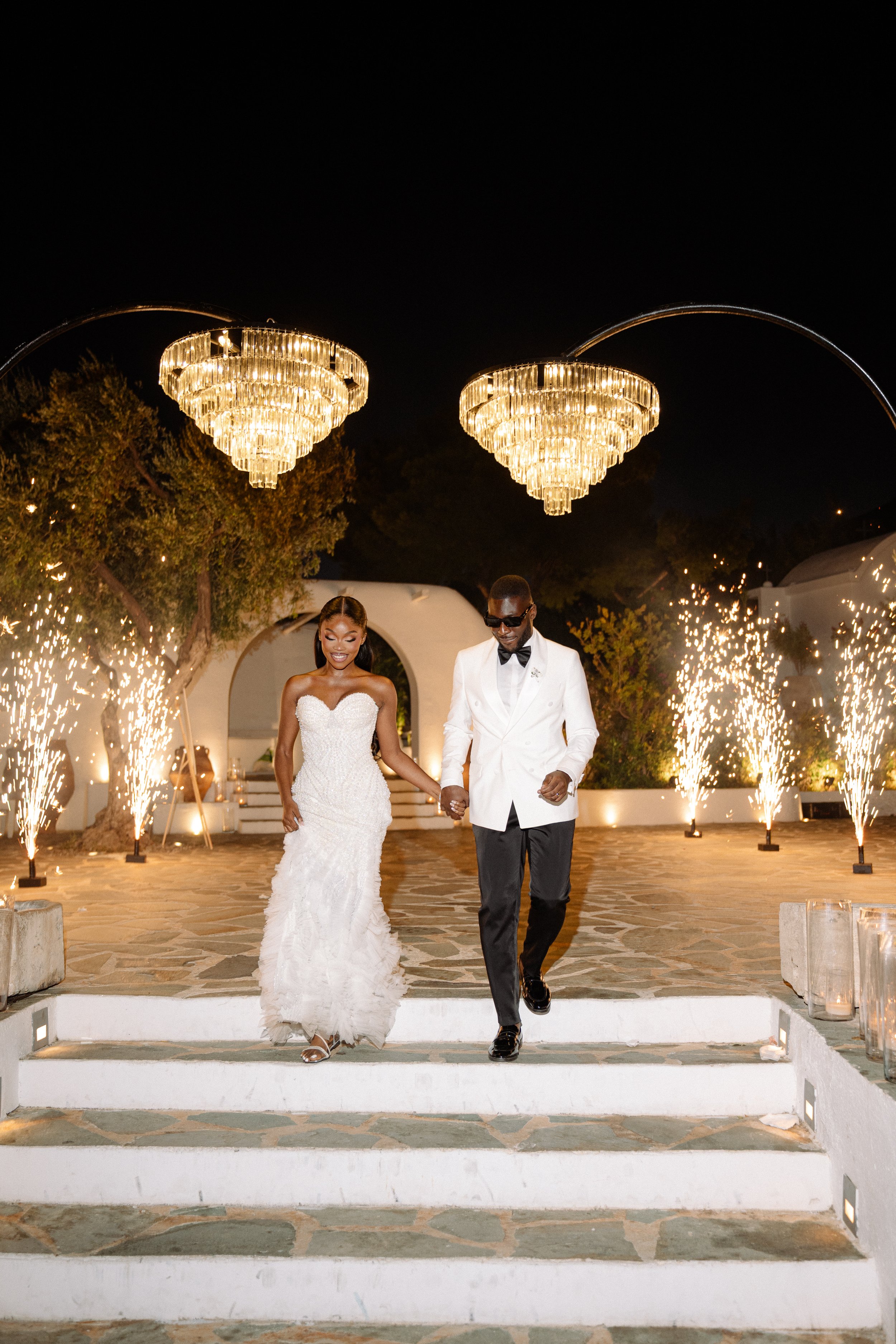 A bride and groom walking down stairs at their wedding reception at night, with fireworks and chandeliers in the background.