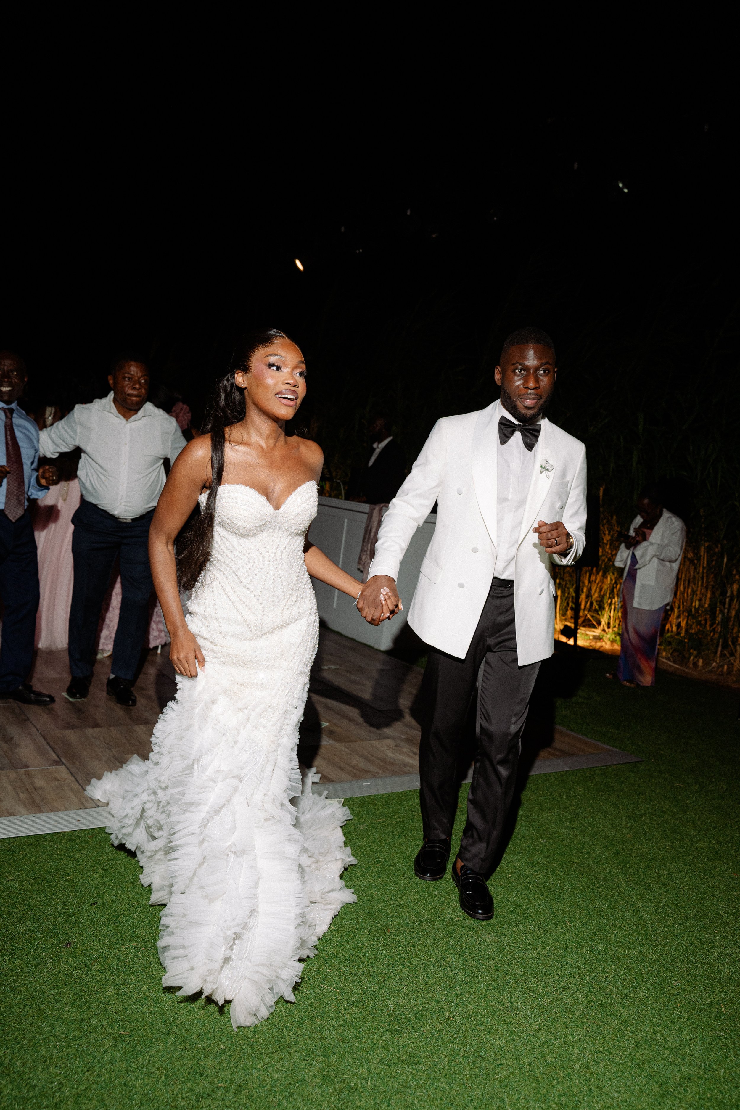 A bride and groom holding hands and dancing at their wedding reception during the night, with wedding guests in the background.