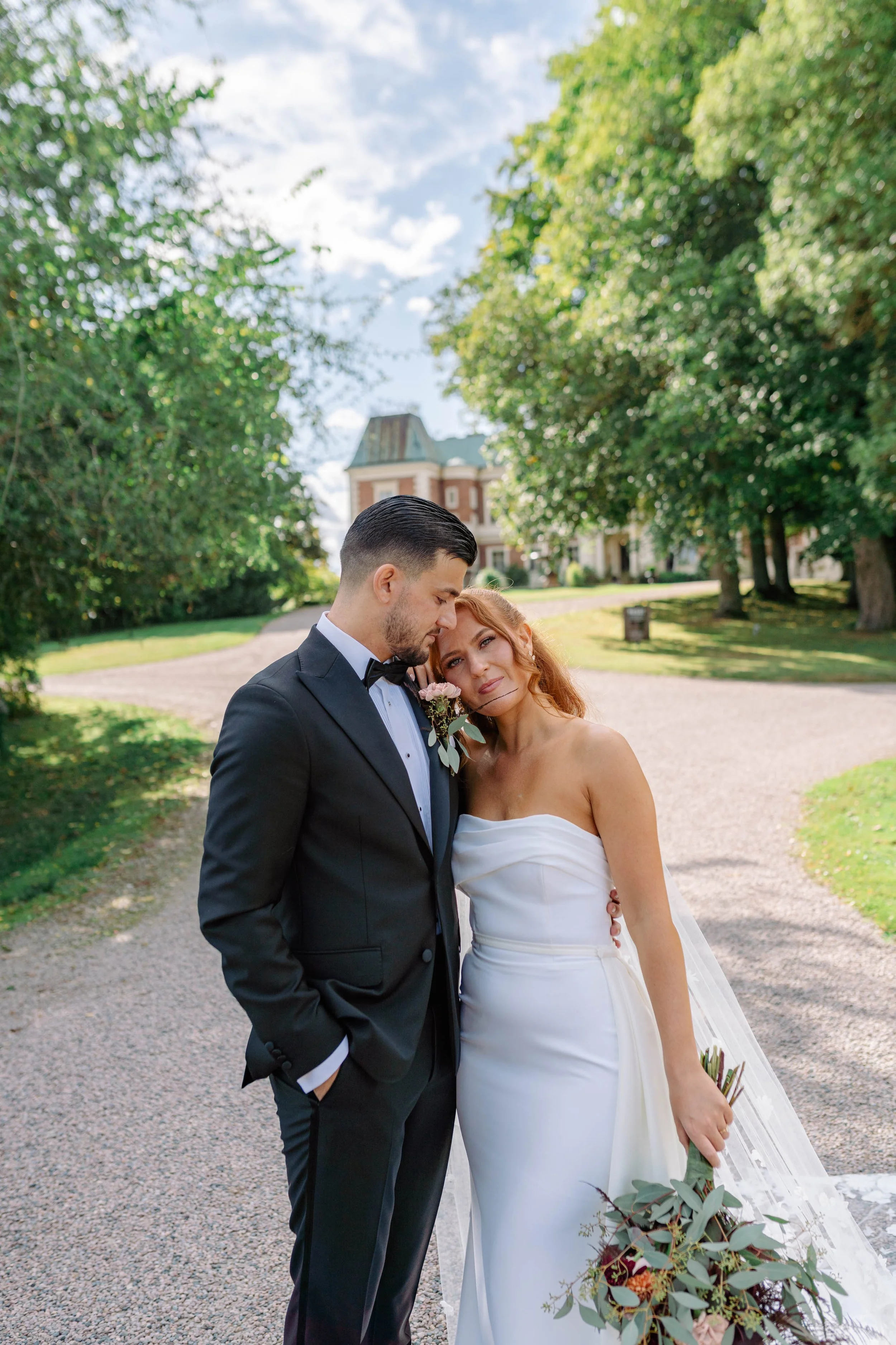 A bride and groom standing close together outdoors on a wedding day, with a historic building and lush greenery in the background.