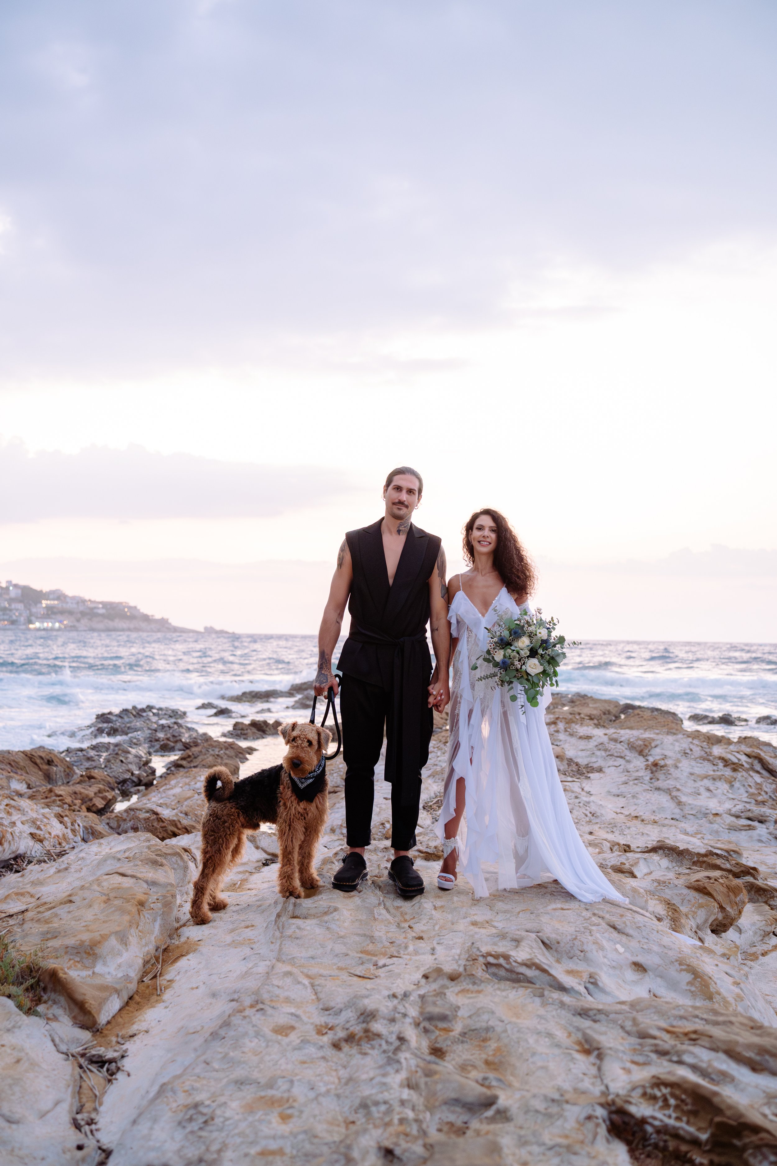 A couple dressed in wedding attire walking hand-in-hand on a rocky beach with their dog, with ocean waves and a cloudy sky in the background.