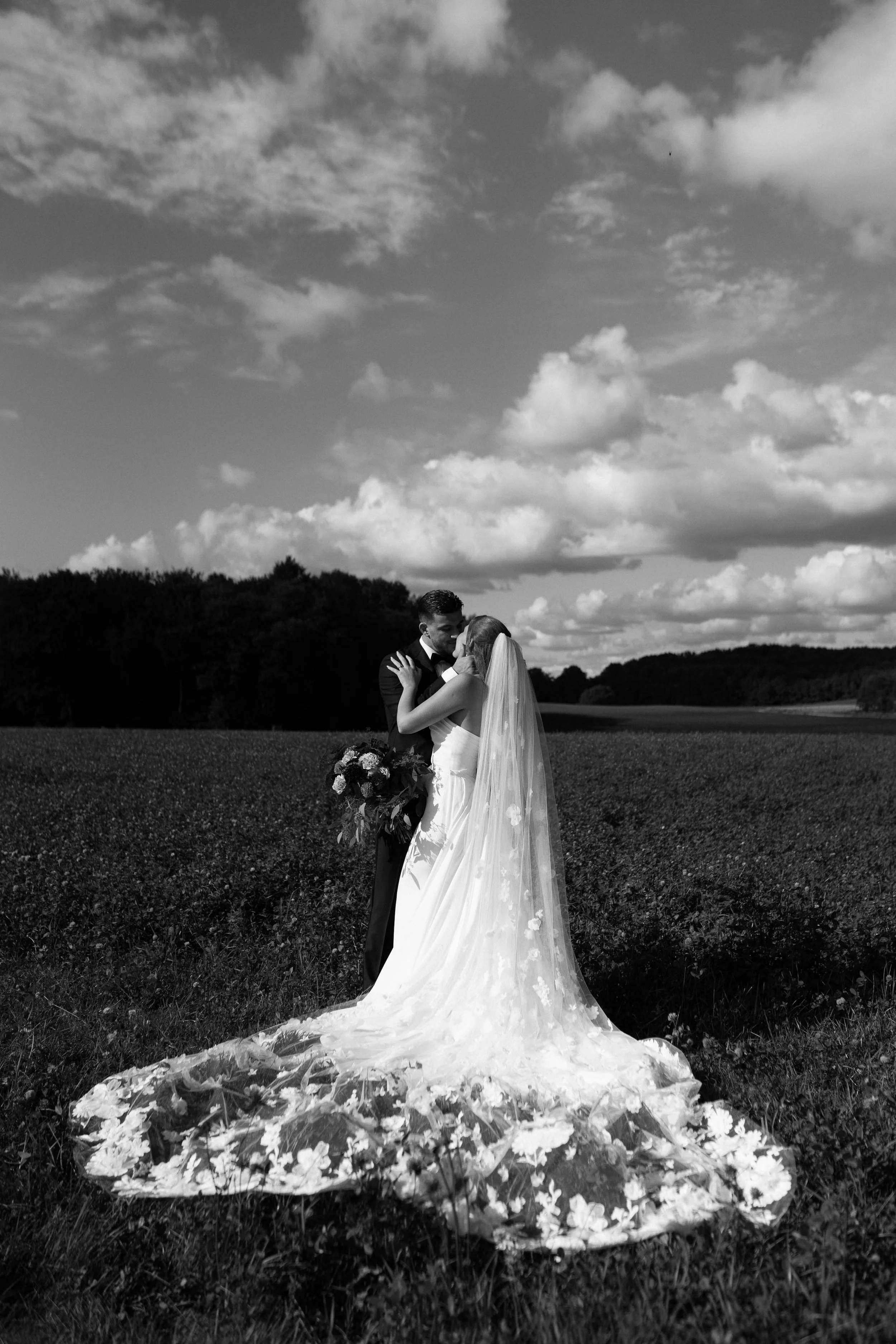 A bride and groom in wedding attire sharing a romantic embrace outdoors with a cloudy sky and trees in the background.