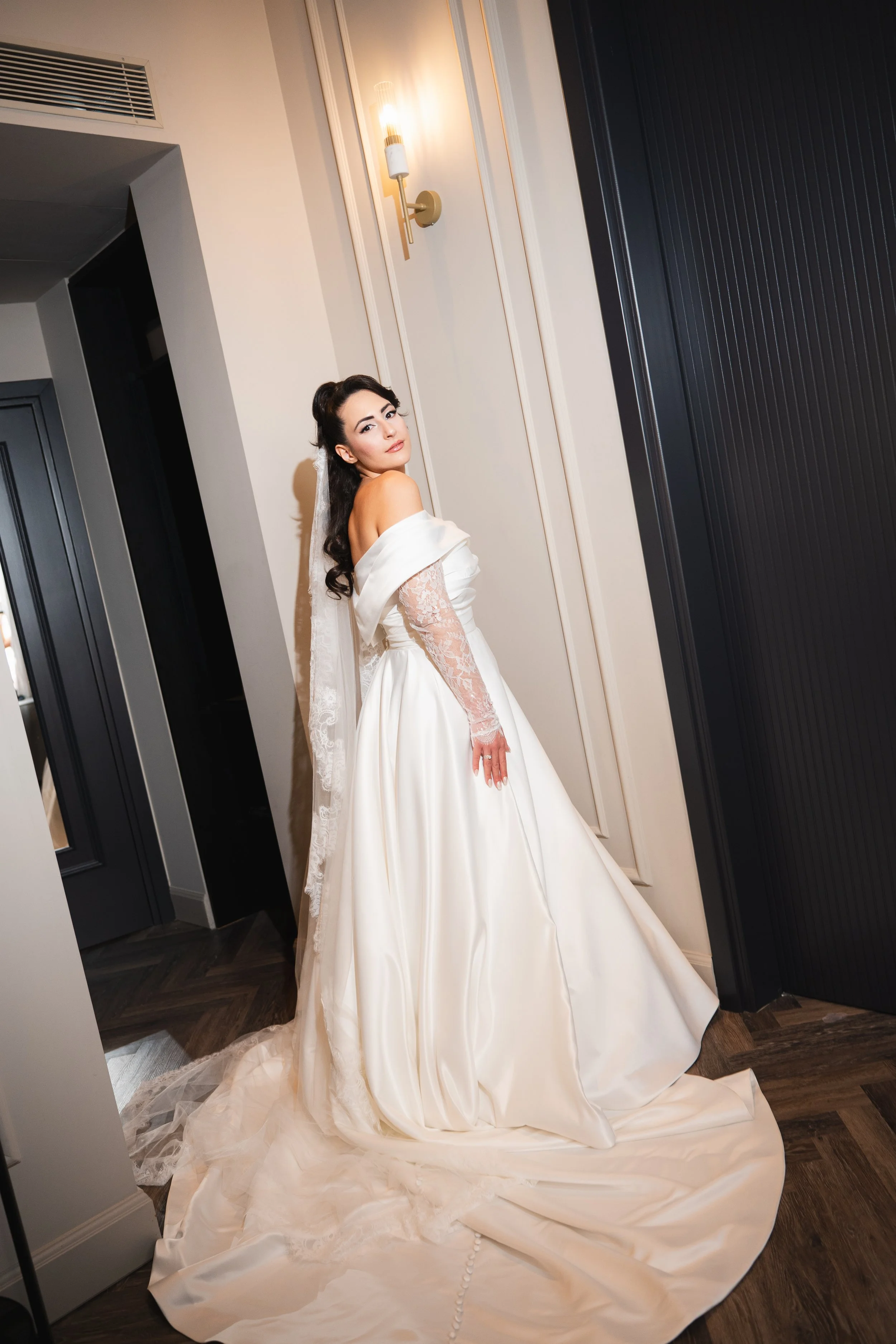 A bride in a white wedding gown with off-the-shoulder lace sleeves and a flowing satin skirt, standing in a modern interior with dark wood flooring and light-colored walls.