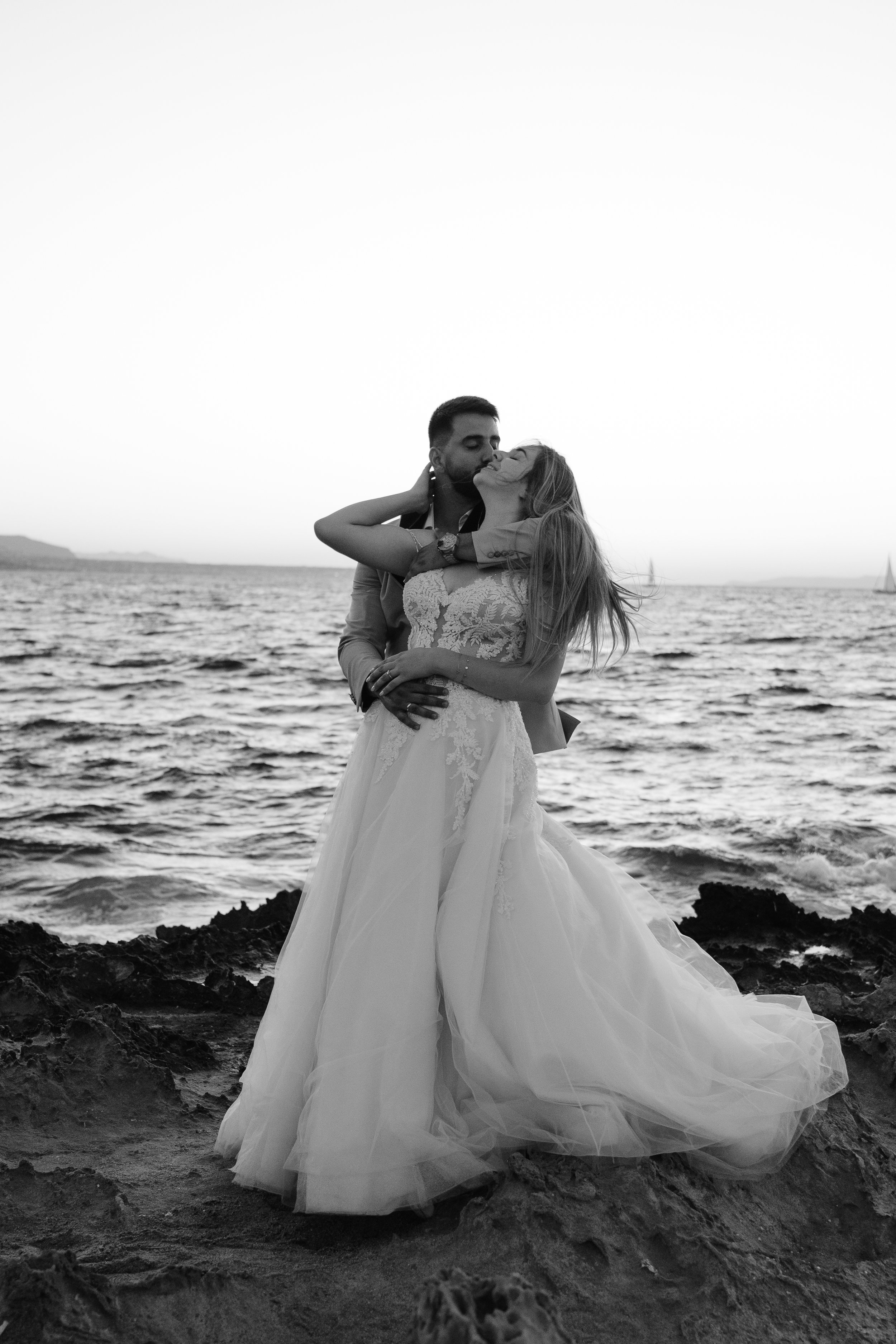 A couple in wedding attire sharing an intimate moment on the rocky shoreline by the sea during sunset, with the woman in a lace wedding dress and the man in a suit.