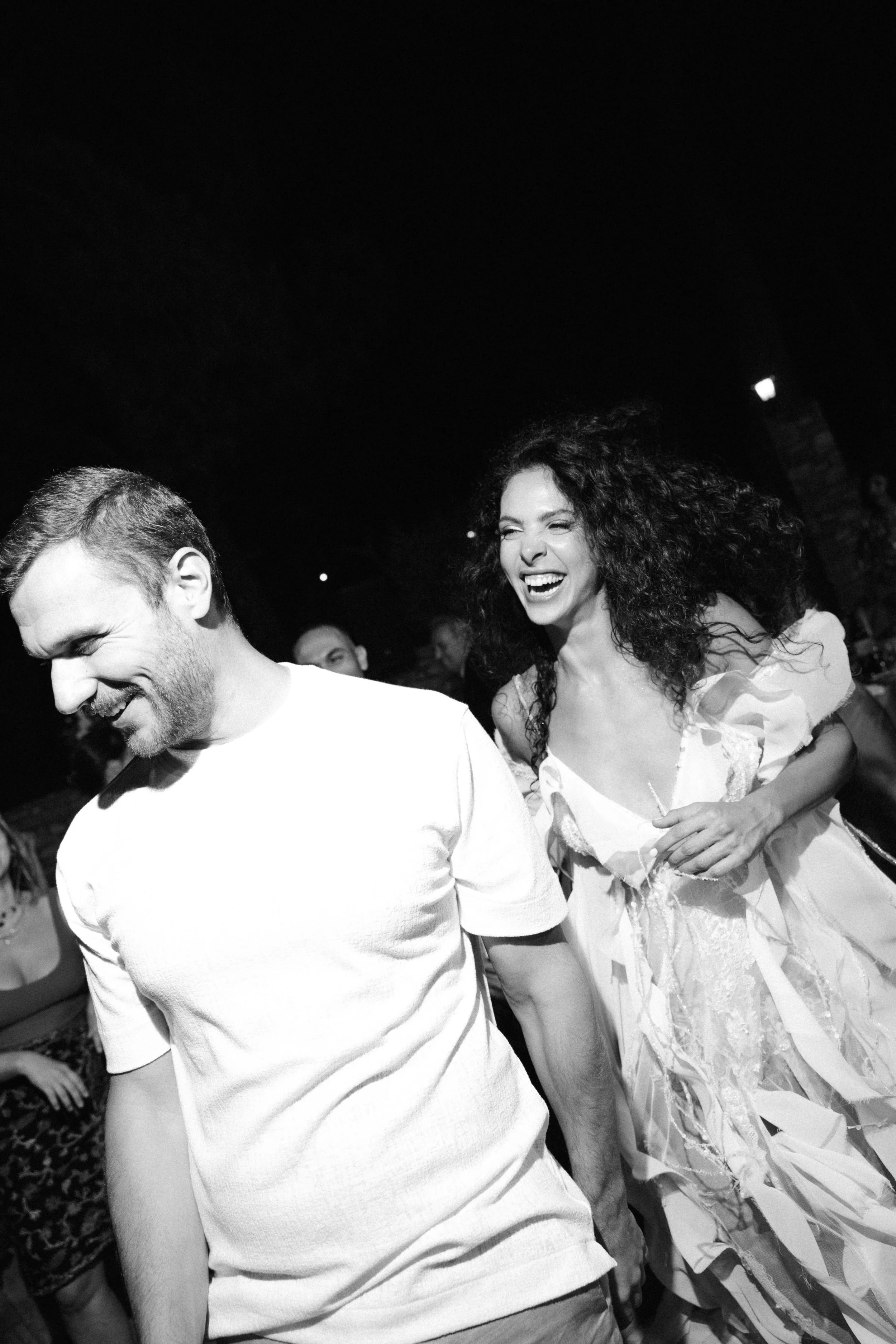 Two people smiling and laughing at a social event, with a woman wearing a light-colored dress with ruffled sleeves and curly hair, and a man in a white T-shirt, against a dark background with other people visible.