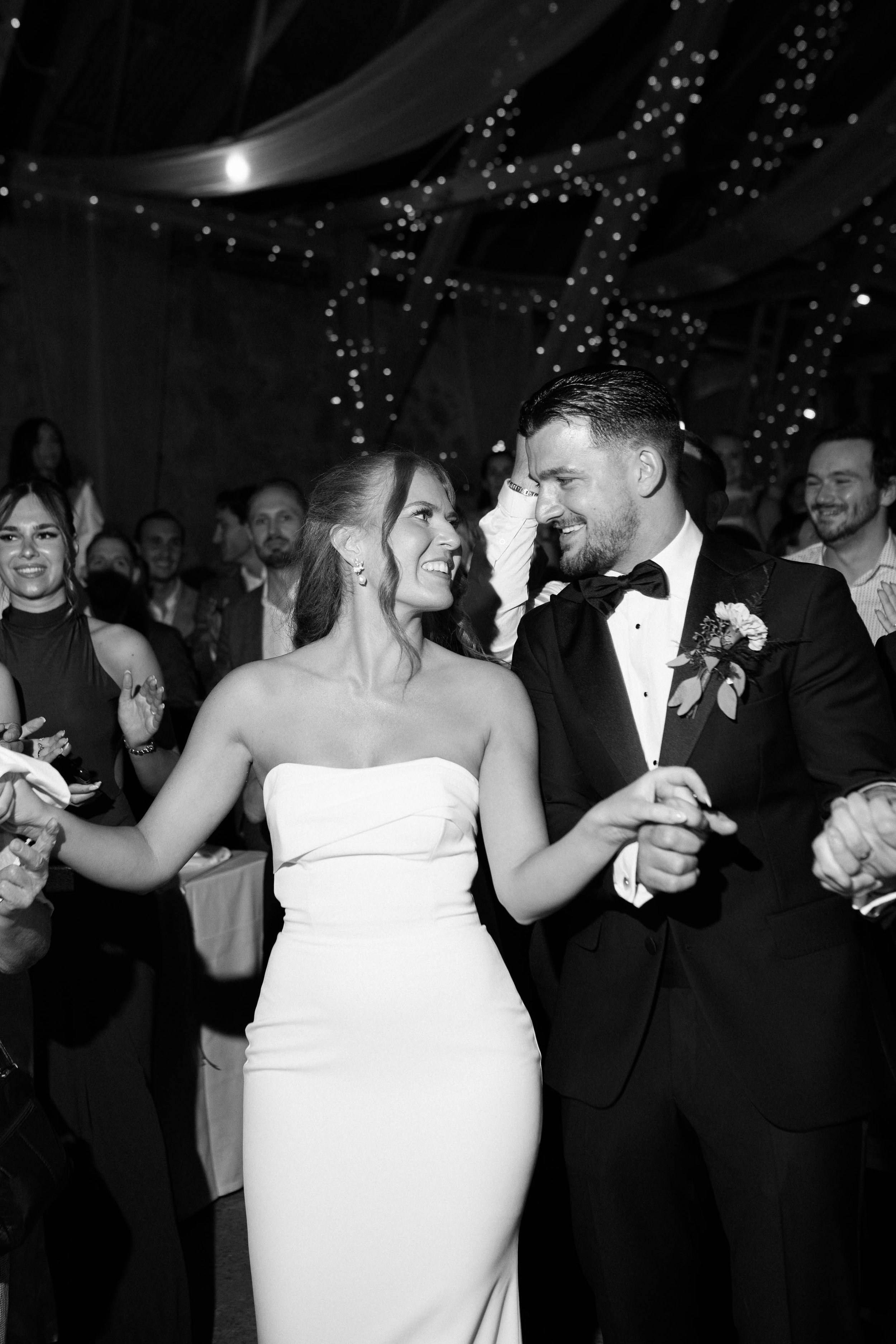Black and white photo of a bride and groom dancing at their wedding reception, smiling at each other, surrounded by friends and family.