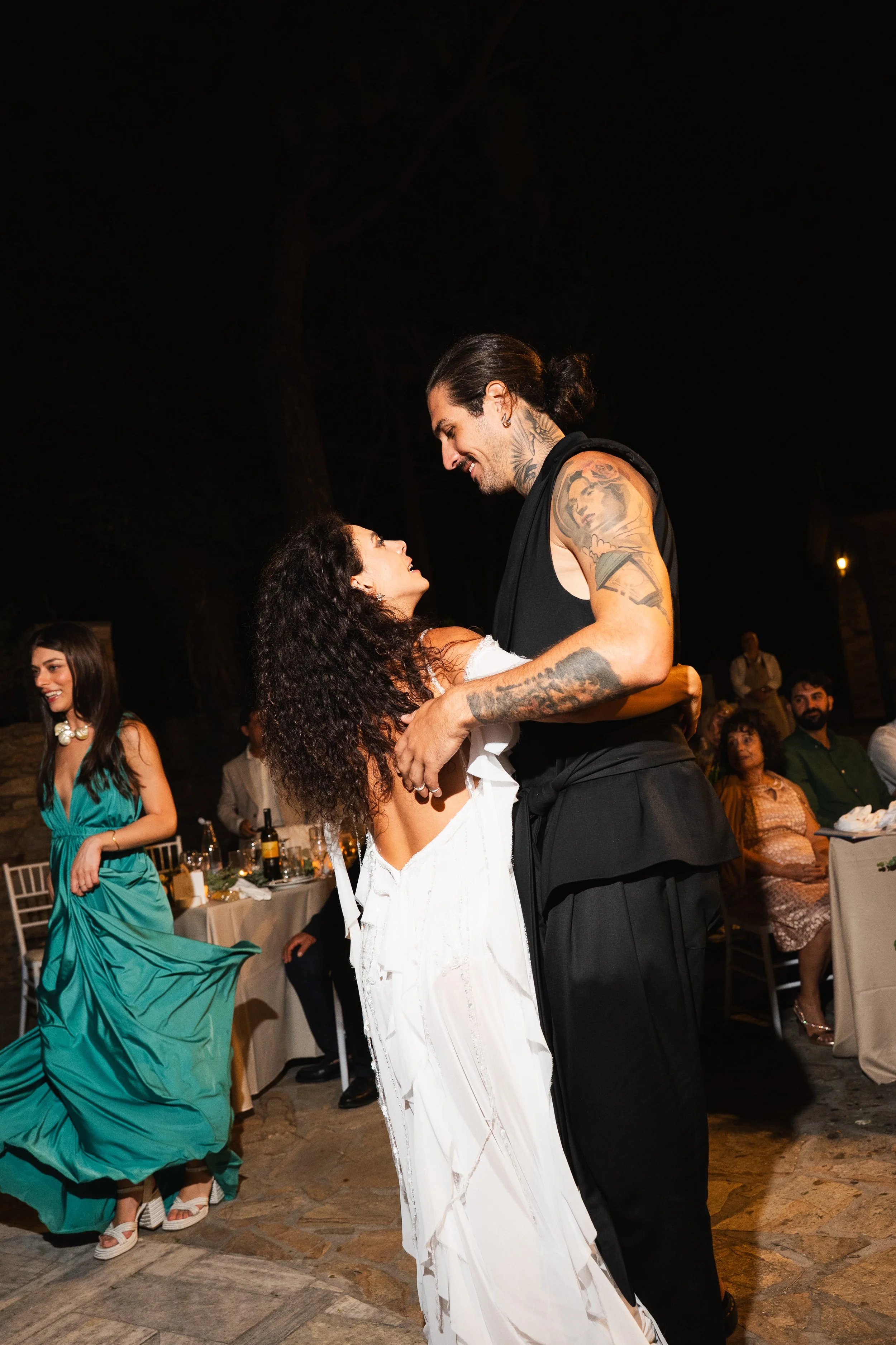 A couple is dancing at a celebration or wedding reception at night, with guests sitting at tables in the background.
