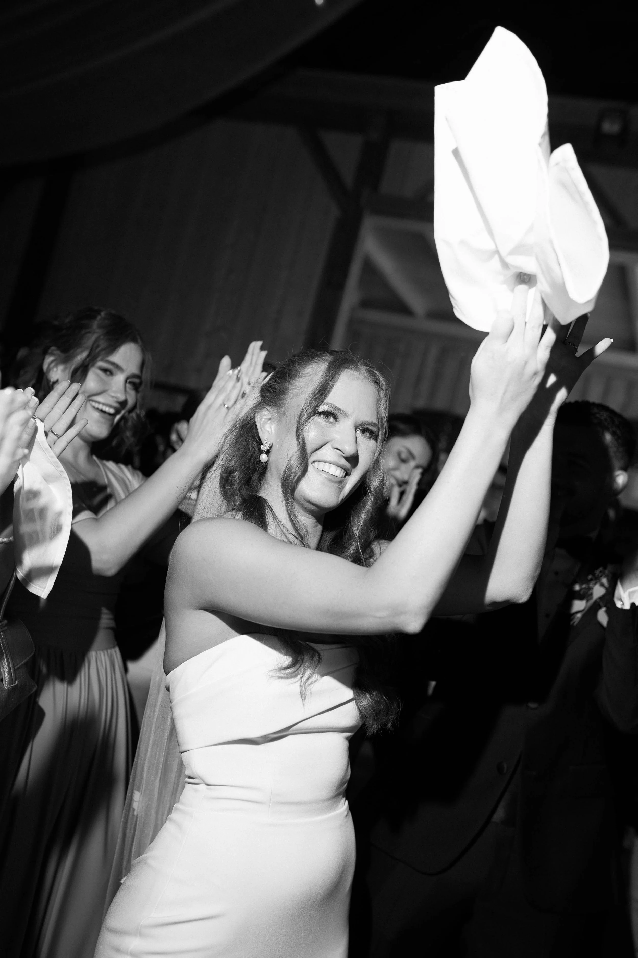 A woman in a strapless wedding dress throws a tissue in celebration at a wedding reception, smiling and surrounded by other smiling guests.