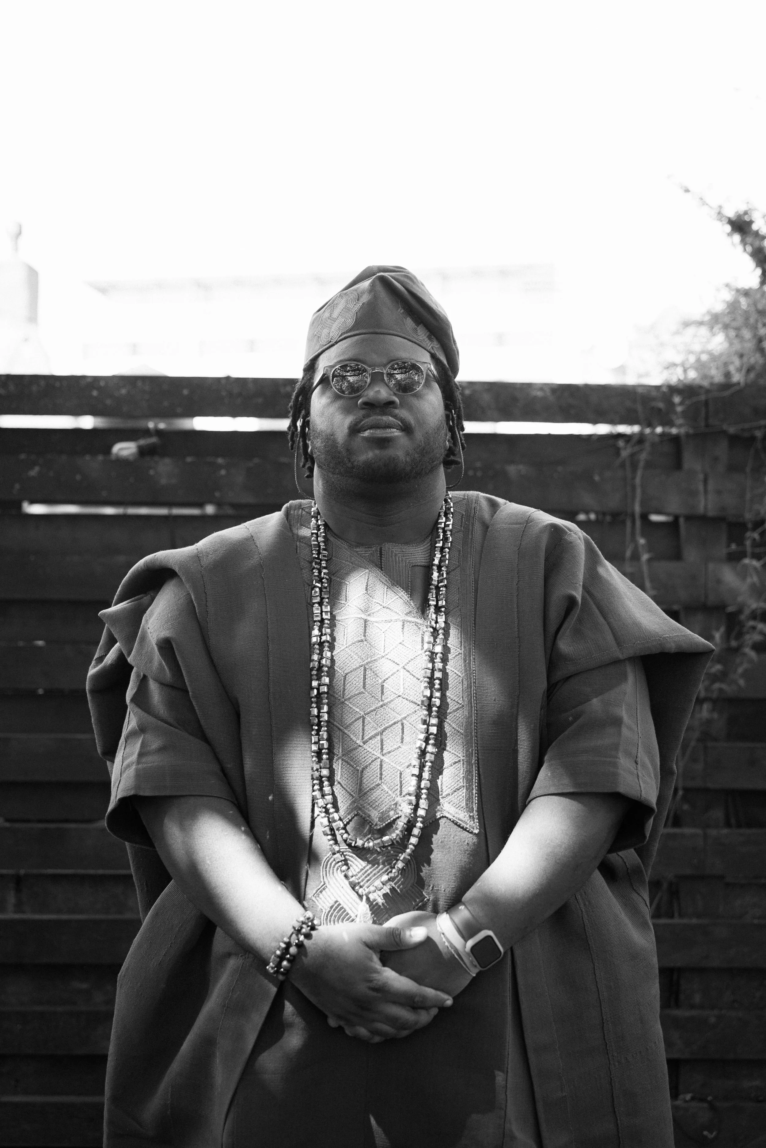 A man with dreadlocks and sunglasses wearing traditional African attire and beaded necklaces, posing outdoors in front of a wooden fence.