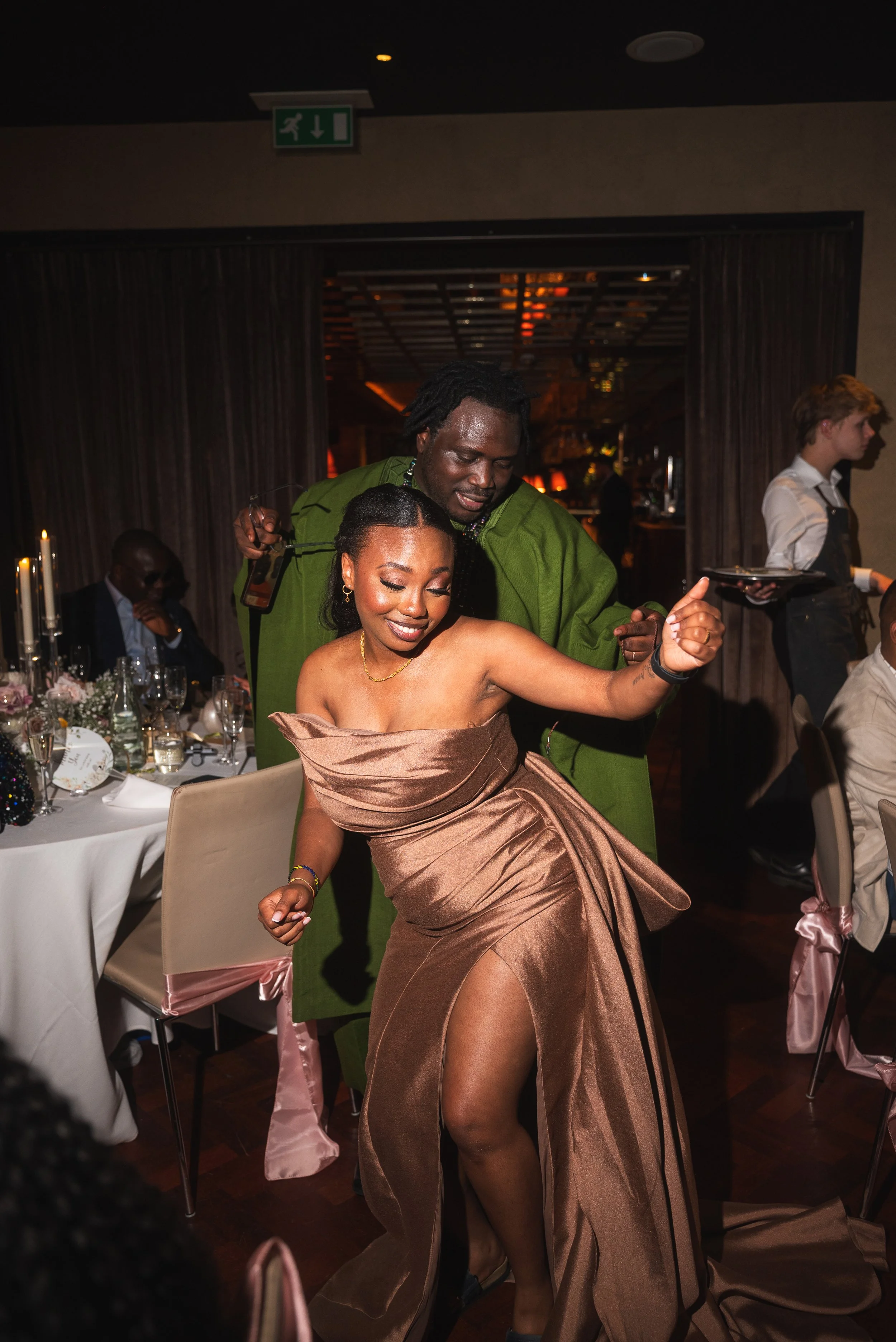People dancing at a formal event with decorated tables and waitstaff in the background.