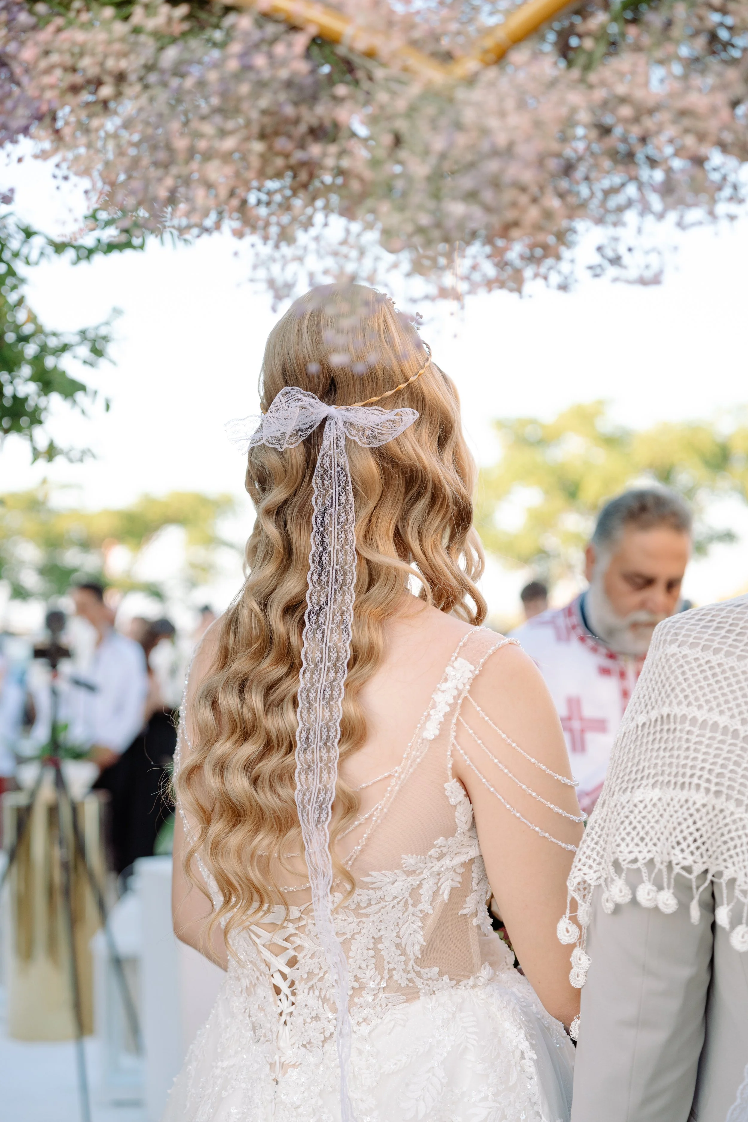A bride with long, wavy blonde hair wearing a white wedding dress with lace details and a lace ribbon in her hair, outdoors during a wedding ceremony.