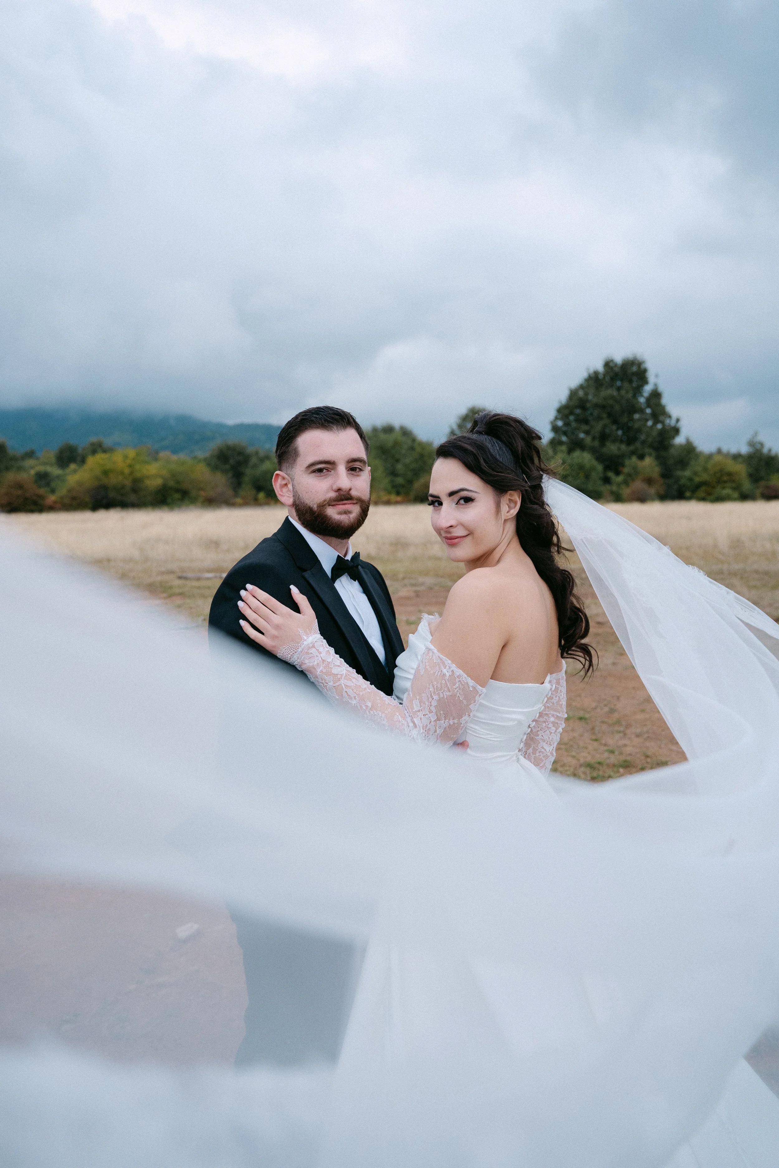 A bride and groom standing outdoors in a field, with mountains and cloudy sky in the background, dressed in wedding attire.