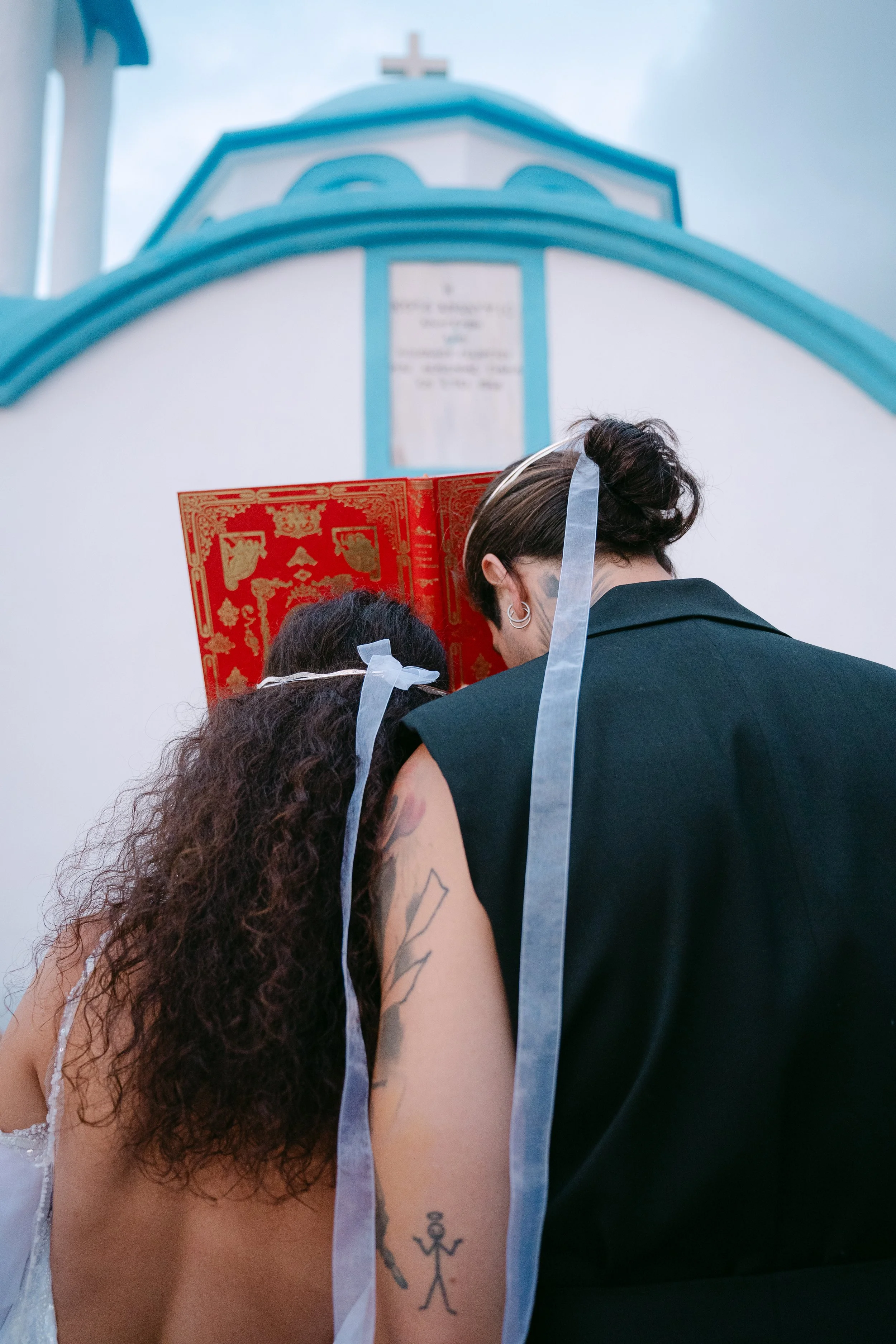 Two people, one with curly hair and tattoo, and another with short dark hair and earrings, standing close with a decorative red book covering faces, in front of a white and blue church.