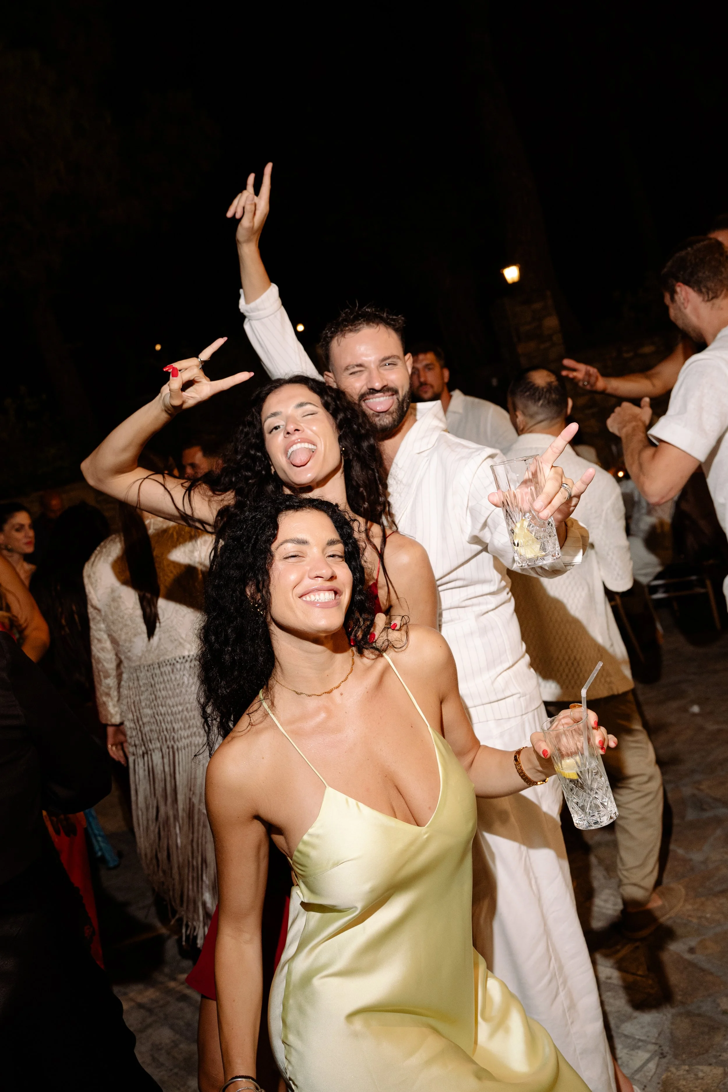 Group of people celebrating at night, smiling, dancing, and holding drinks, with a dark outdoor setting.