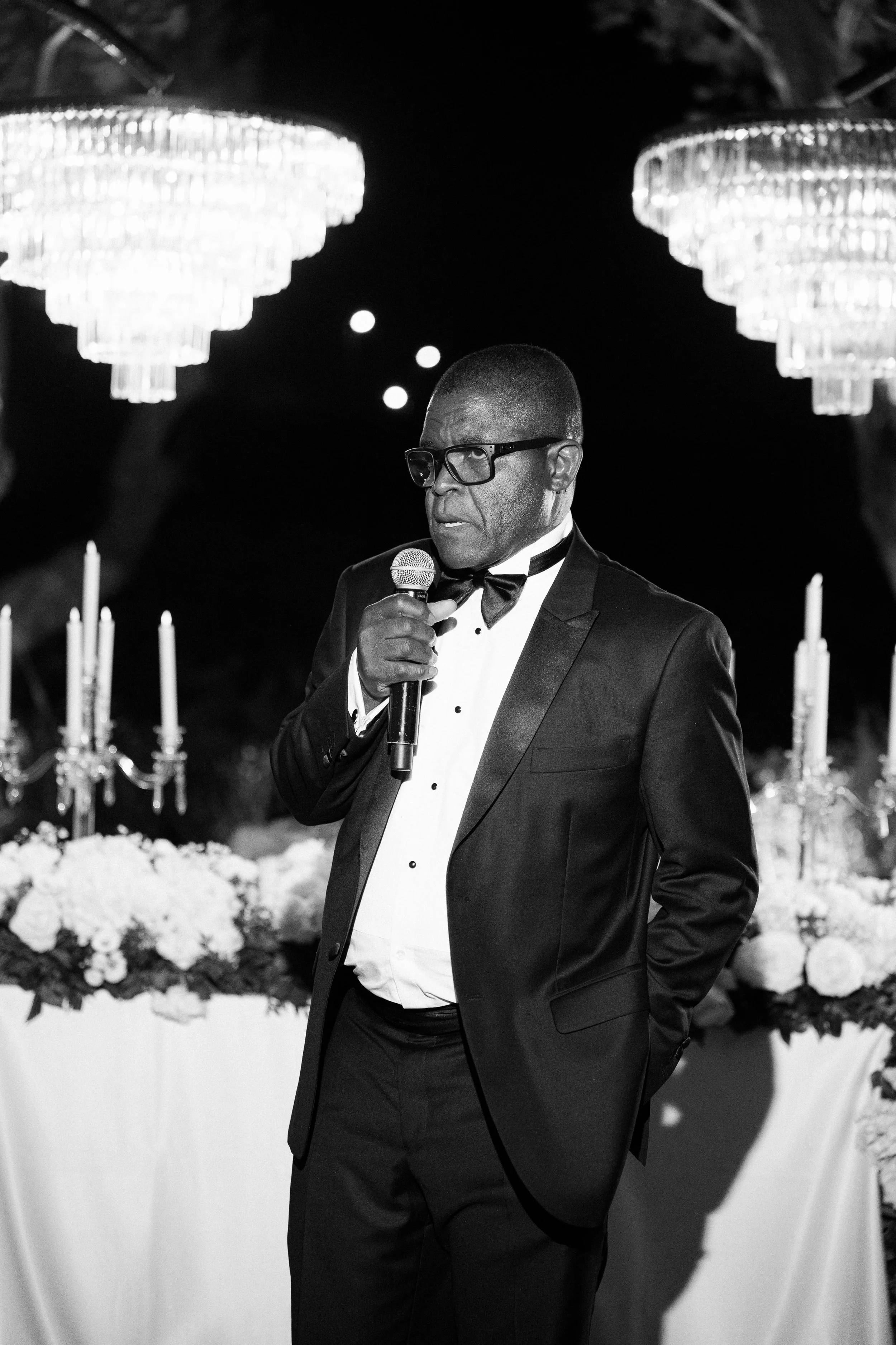A man in a tuxedo and glasses holding a microphone at a formal event, with chandeliers and candles in the background.