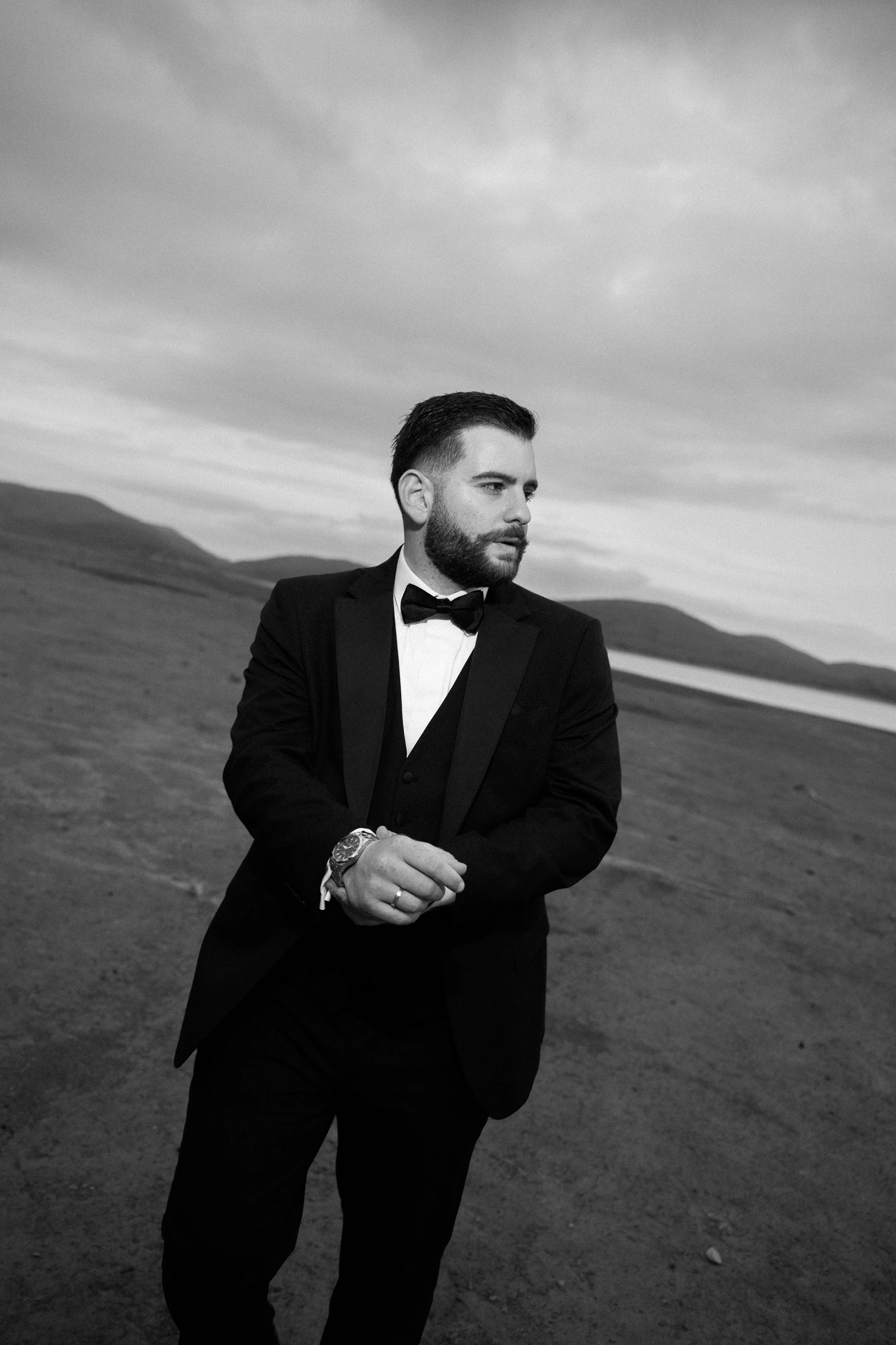 A man in a tuxedo standing outdoors with a landscape of hills and a body of water in the background, looking to his right.