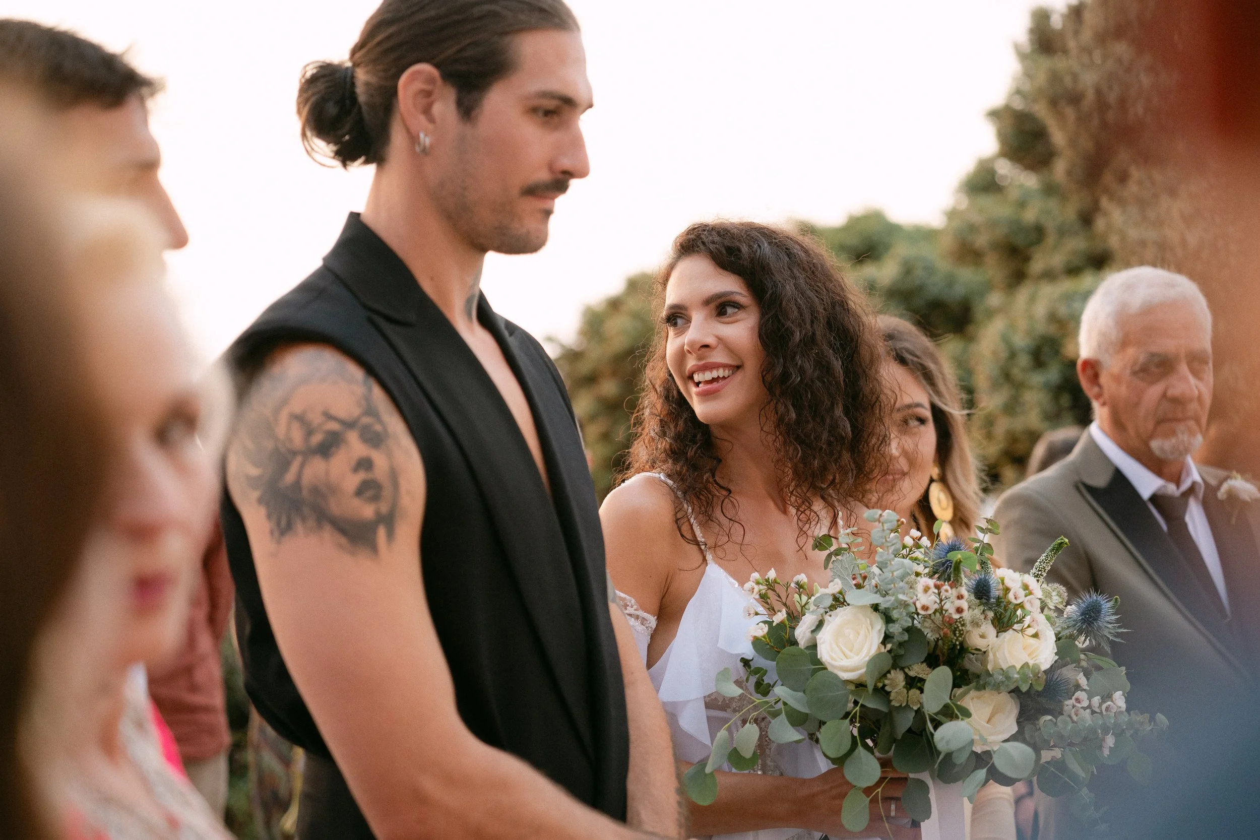 A woman in a white dress holding a bouquet of flowers, smiling at a man with tattoos on his arm, at an outdoor wedding ceremony.