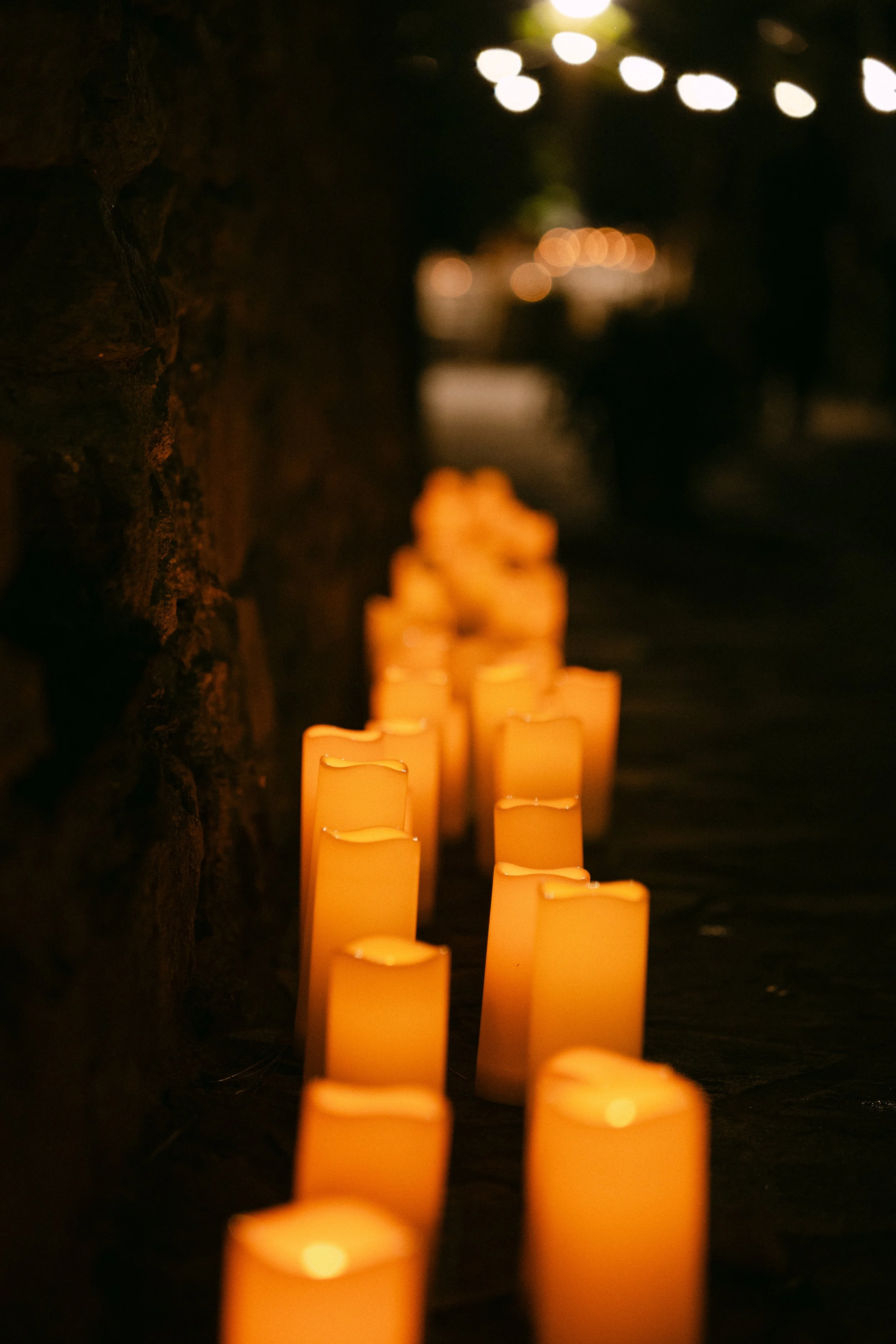 A row of glowing candles aligned on the ground near a stone wall at night, with blurred lights and shadows in the background.