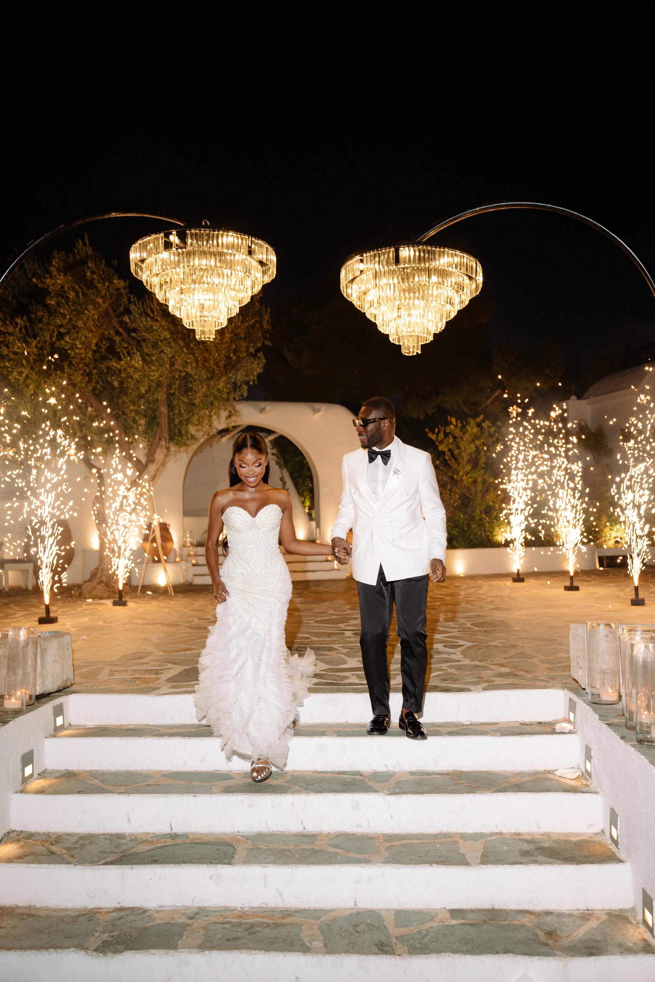 A bride and groom walking down the stairs during their wedding celebration at night, with sparklers and lights in the background.