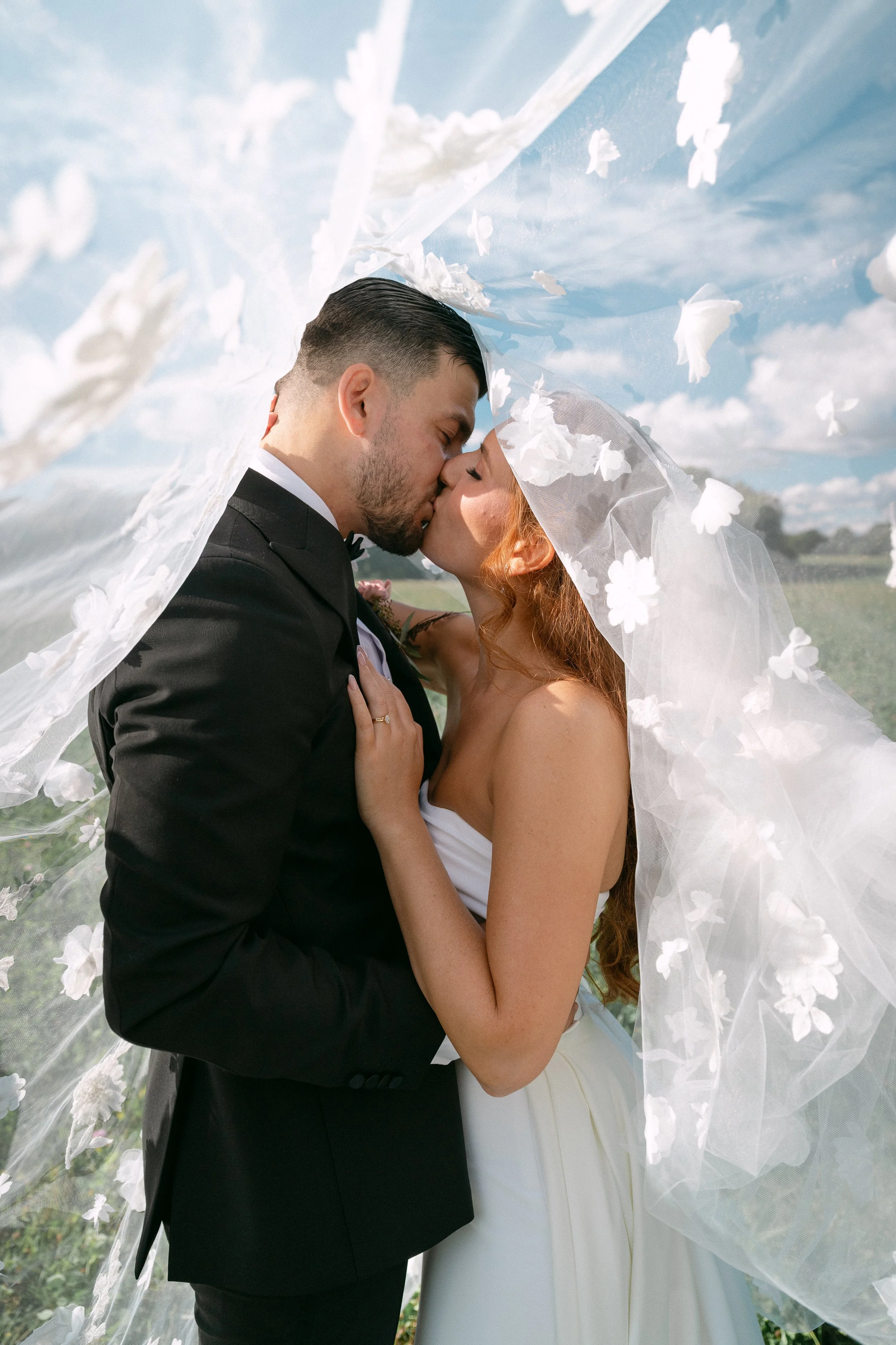 A bride and groom sharing a kiss under a veil with floral details during their outdoor wedding.