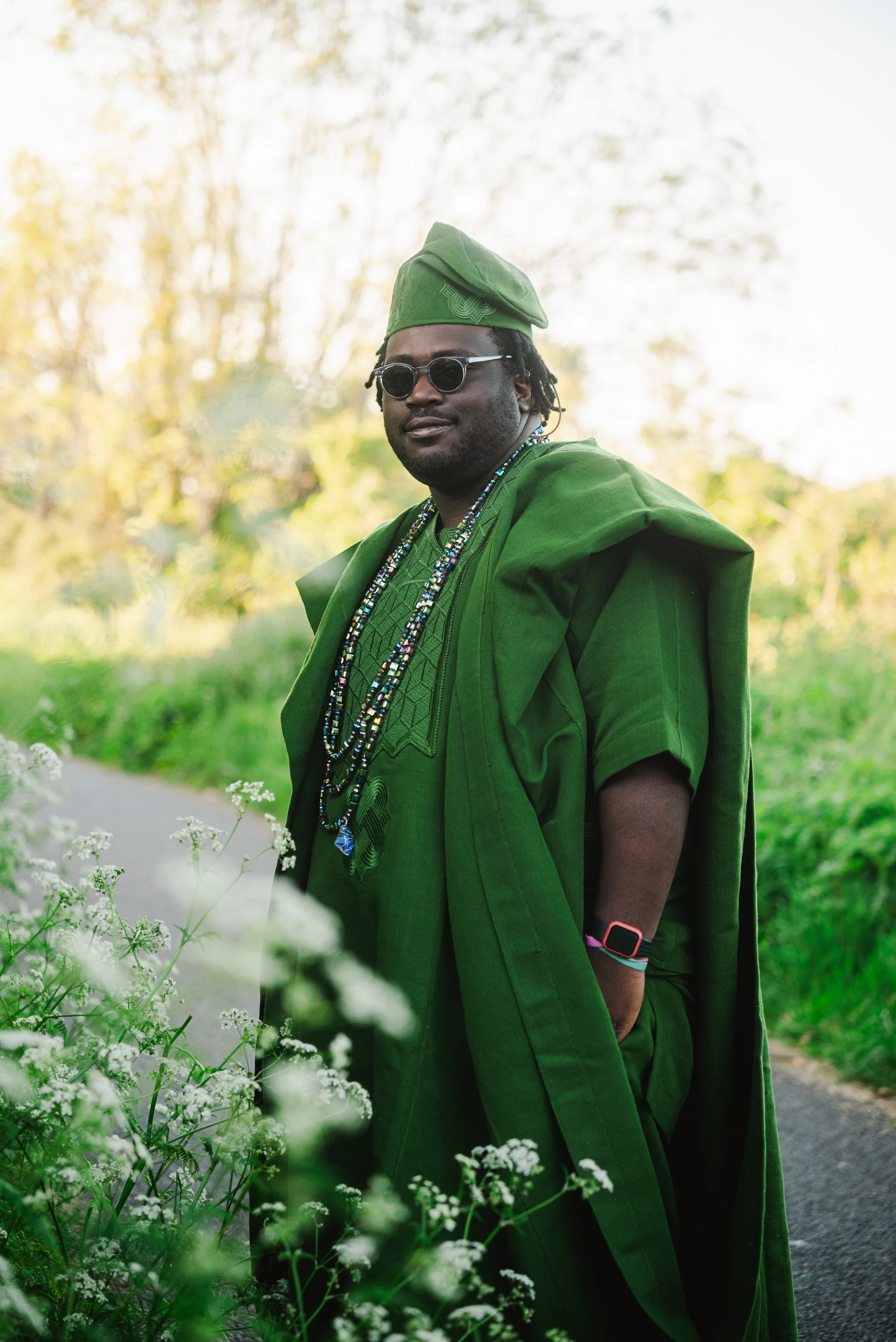A man dressed in all green traditional clothing stands outdoors with a blurred green background, wearing sunglasses, a green cap, and colorful beaded necklaces.