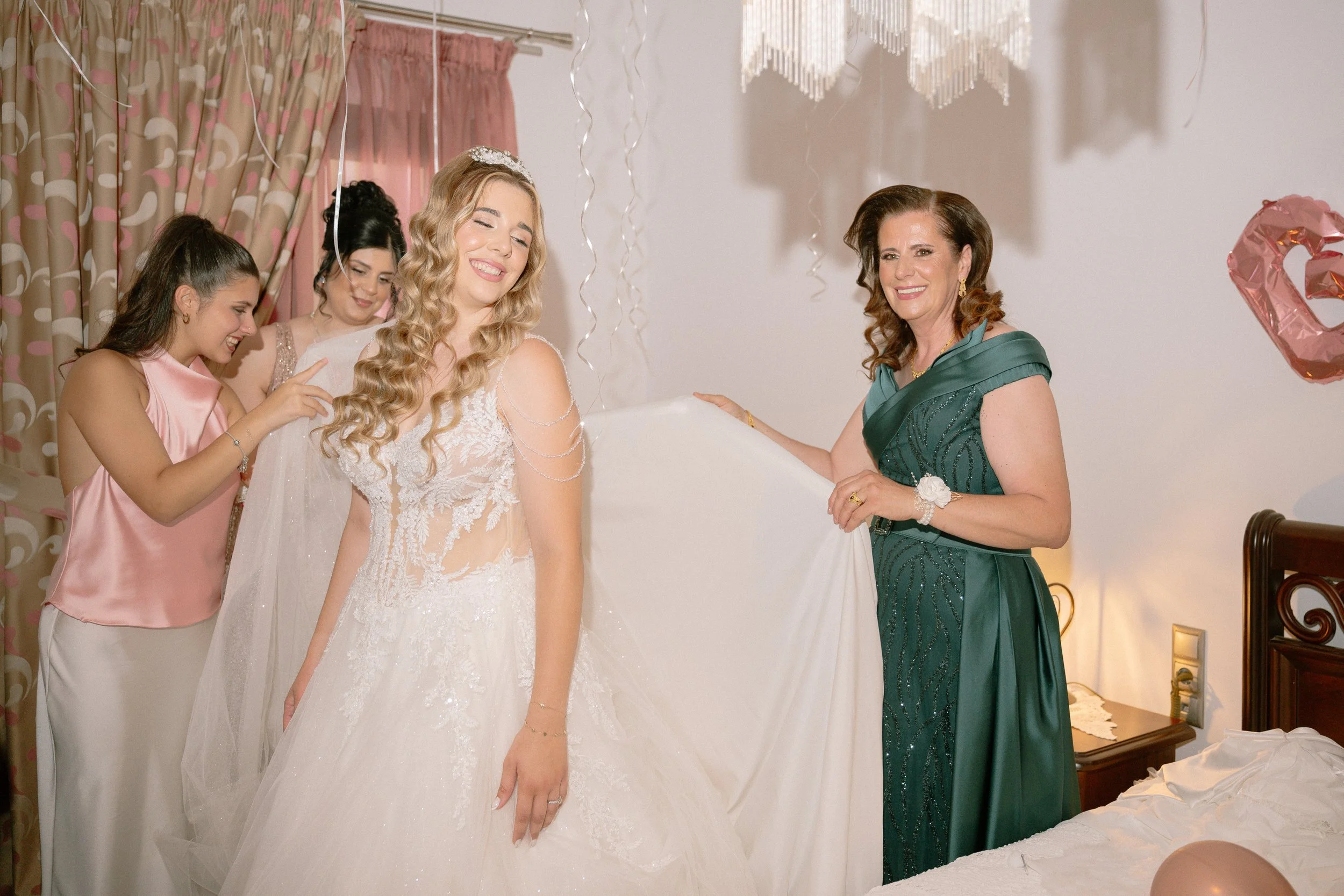 A bride in a white wedding dress smiling with eyes closed as she stands next to a woman in a dark green dress, with three other women nearby, all in a decorated room with pink and beige curtains. Decorations include balloons and hanging ornaments.