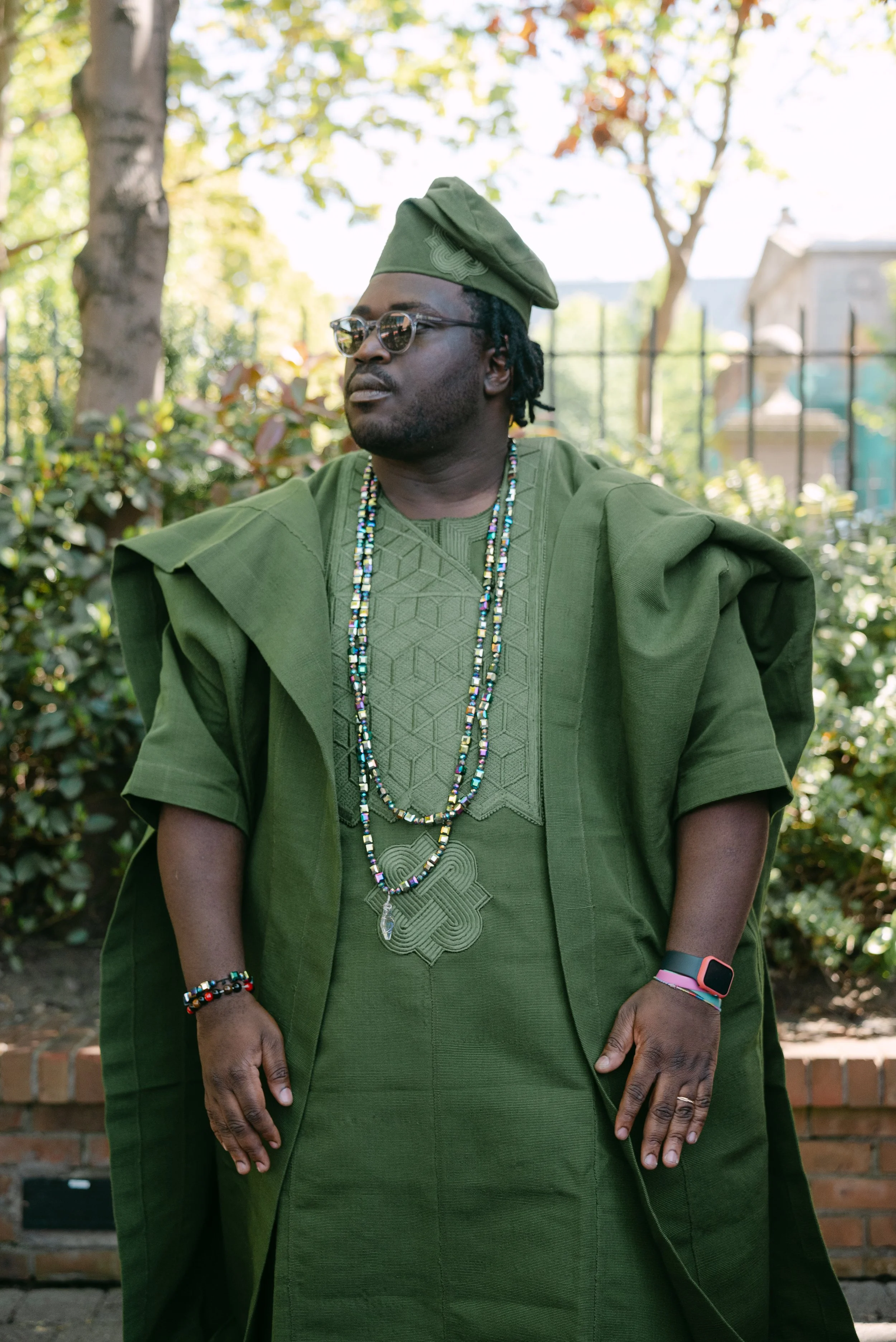 A man dressed in traditional African clothing in shades of green, wearing multiple beaded necklaces, glasses, and a green cap, standing outdoors in front of trees and a fence.