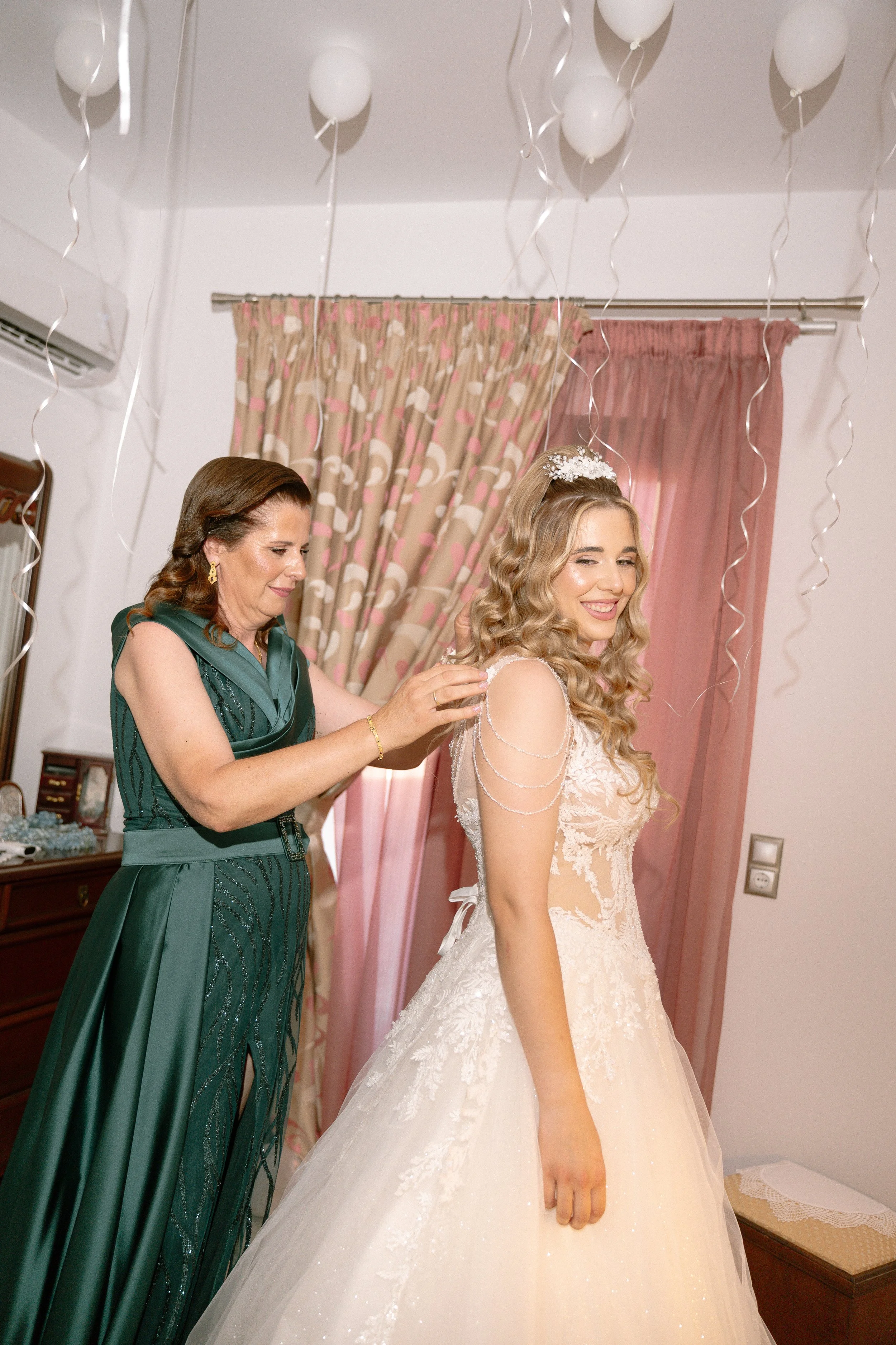 A bride getting ready in a bedroom with a woman helping her, decorations include white balloons and pink curtains.