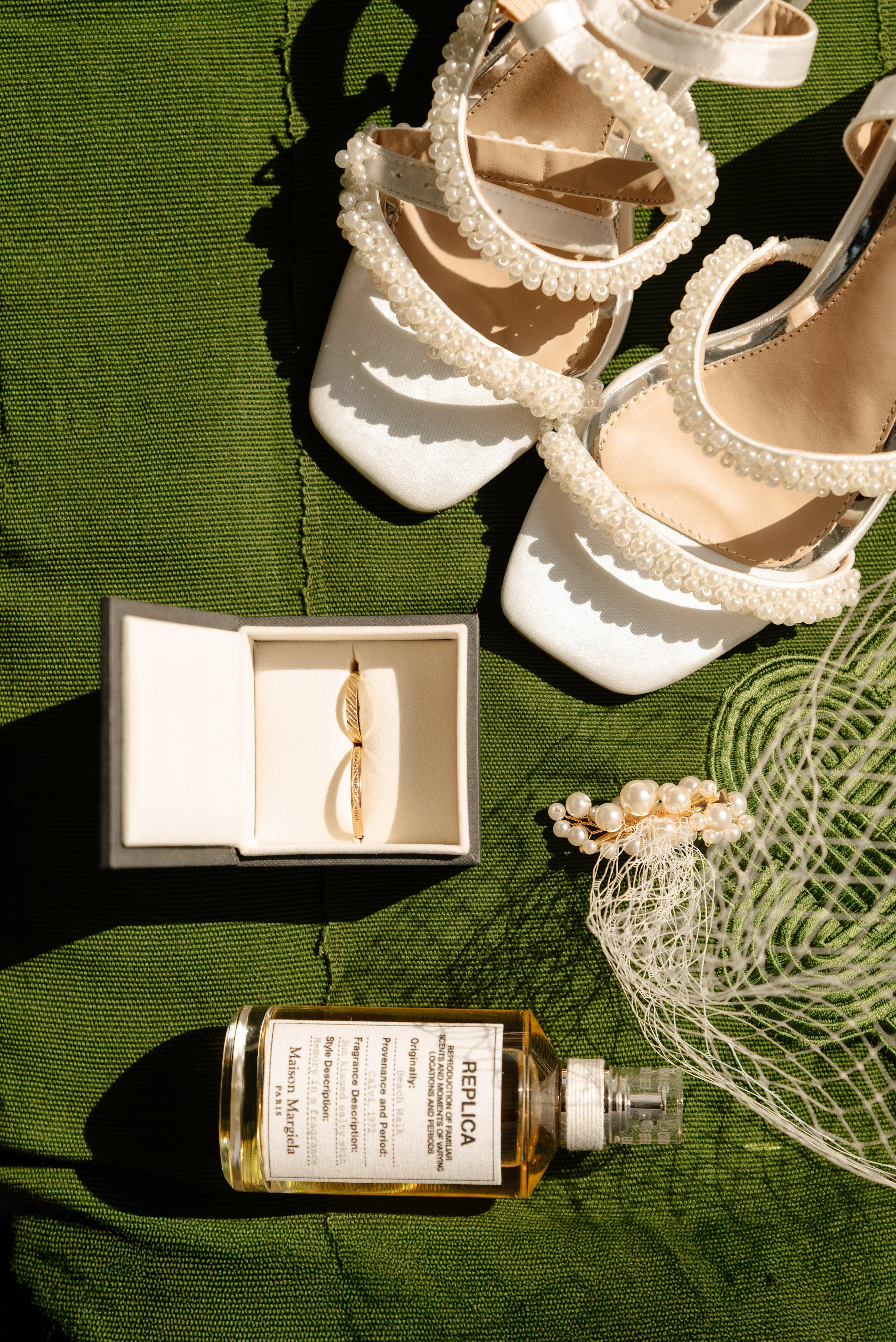 A pair of white high-heeled sandals decorated with pearl embellishments, a small jewelry box with a gold ring inside, a pearl bracelet, a bottle of perfume, and a decorative white mesh accessory are arranged on a green textured fabric surface.