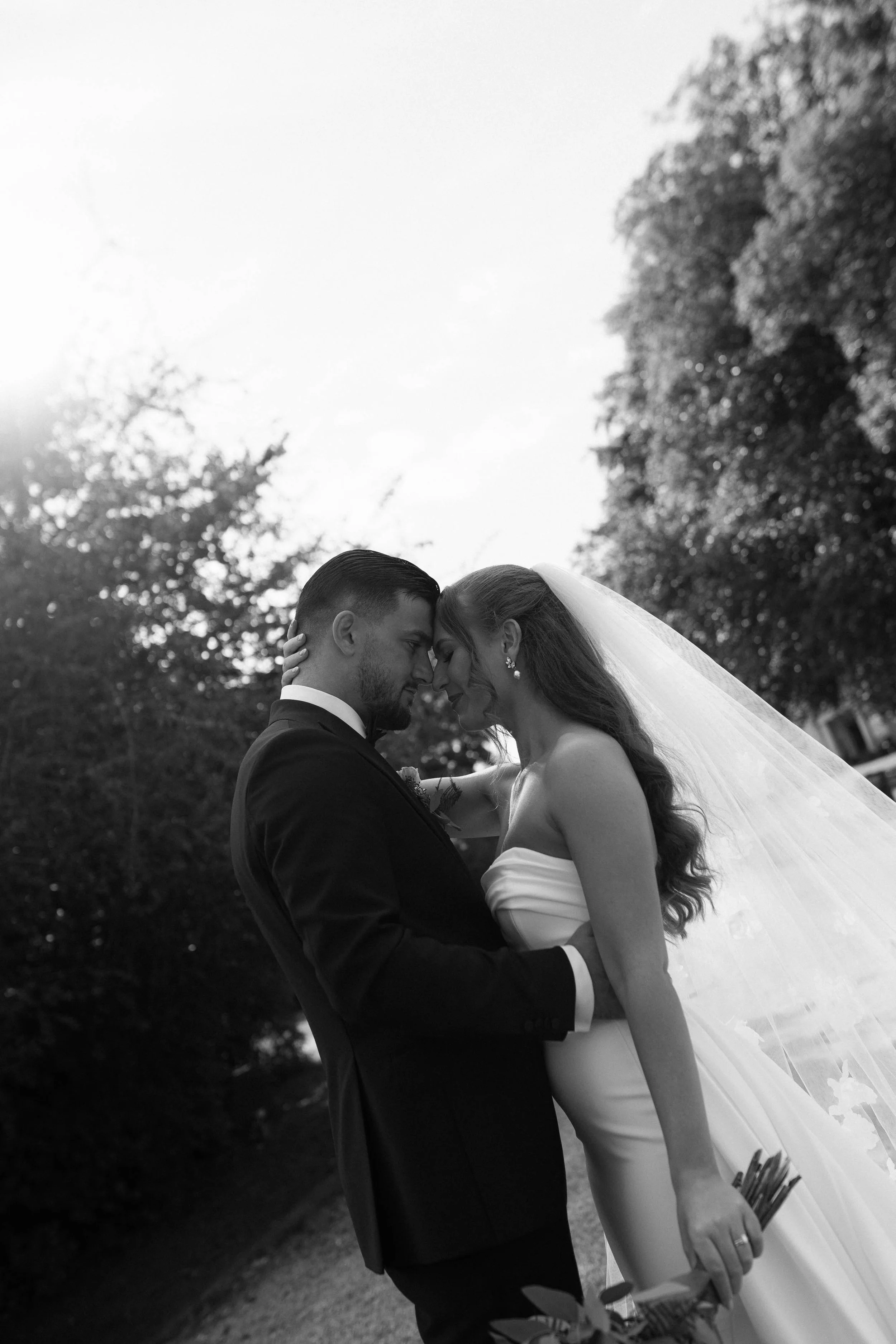 Black and white photo of a bride and groom in an outdoor setting, touching foreheads with eyes closed, bride holding flowers, groom in a suit, trees in the background.