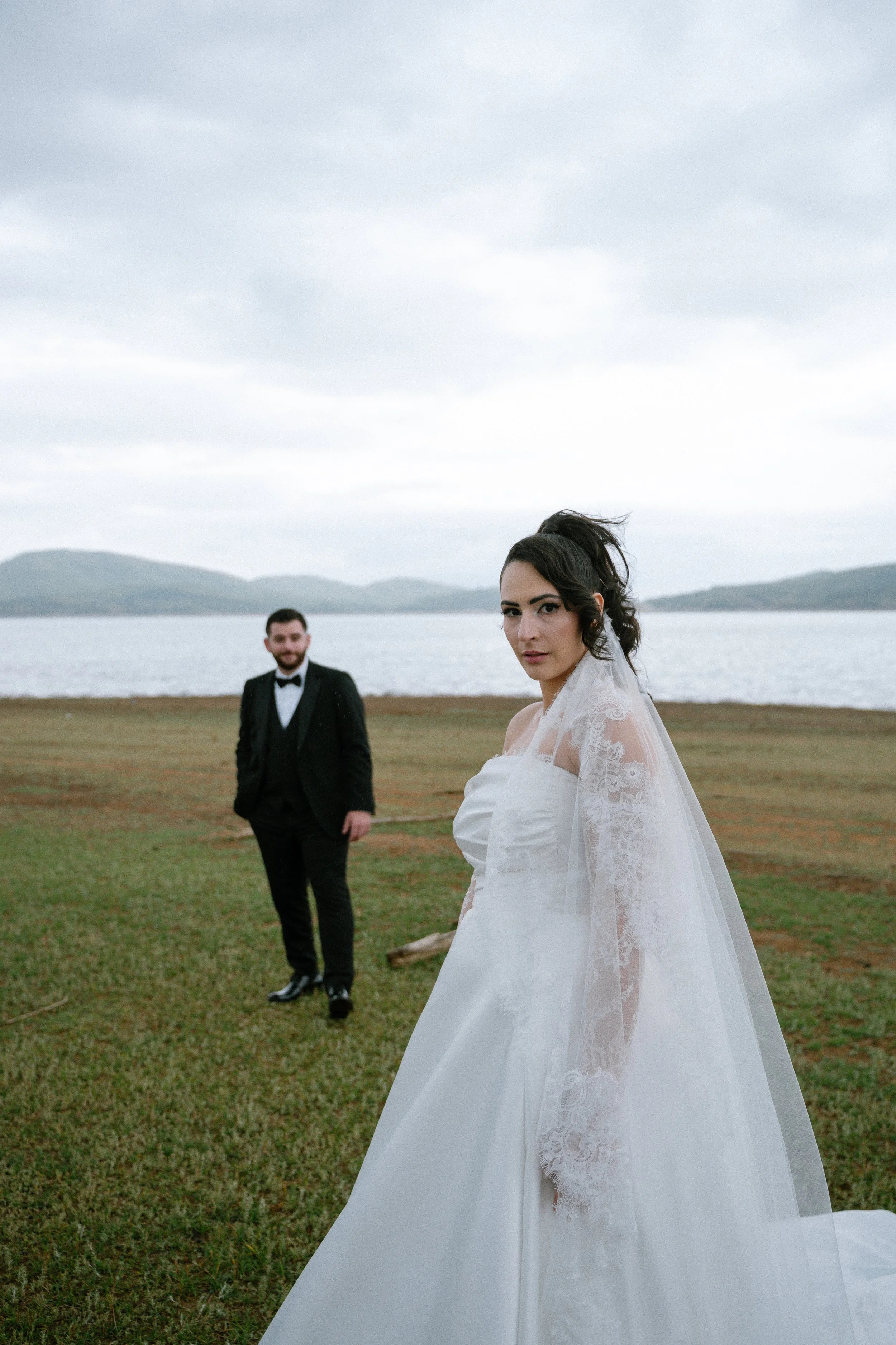 A bride in a white wedding gown with lace details on the sleeves stands on green grass near a body of water, with a groom in a black tuxedo and bow tie in the background, under a cloudy sky.