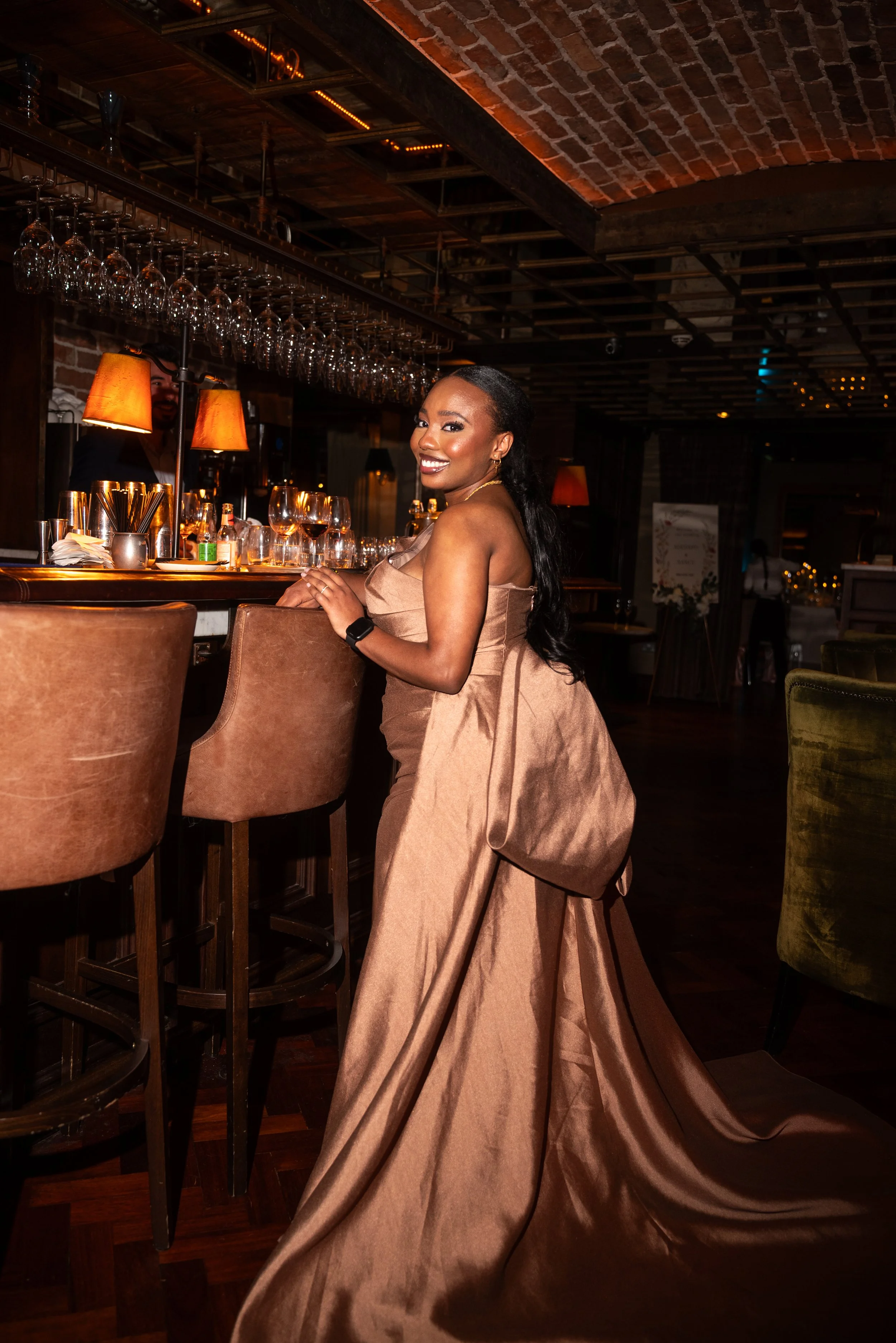 A woman in a tan gown smiling and leaning against a bar in a dimly lit restaurant or bar with glasses, bottles, and warm lighting.