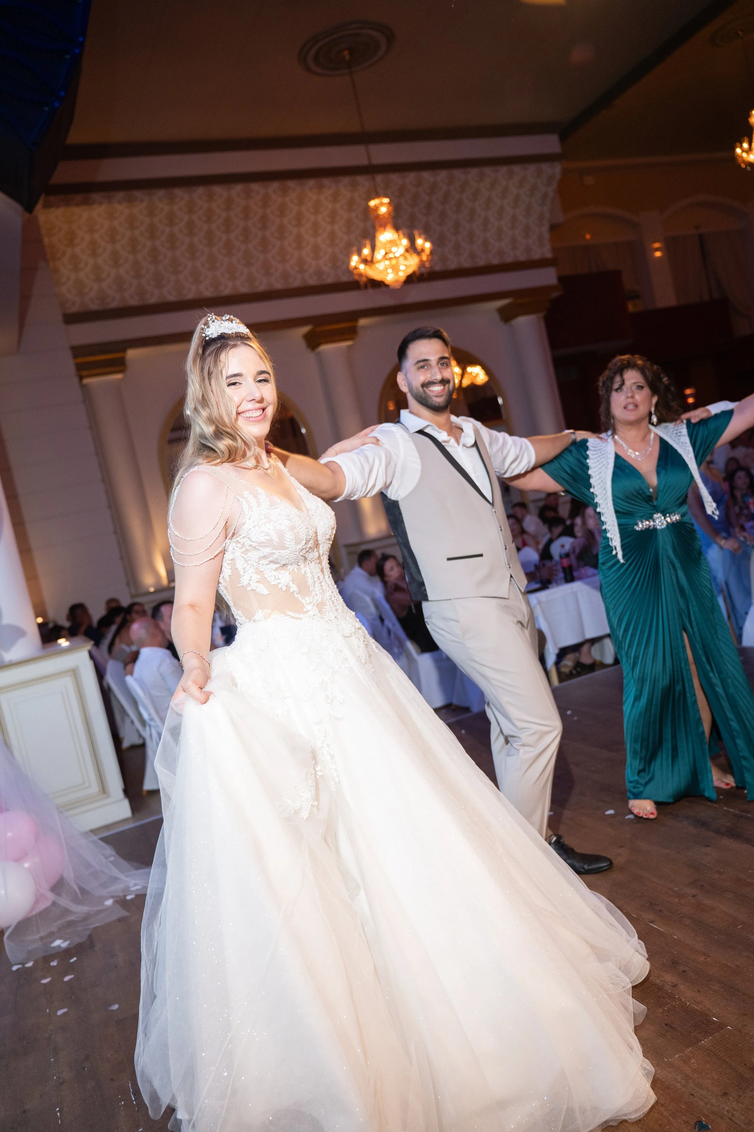 A bride and groom dancing at their wedding reception, smiling, with a woman in a green dress. The bride is in a white wedding gown with lace details, and the groom is in a light tan vest and pants with a white shirt.